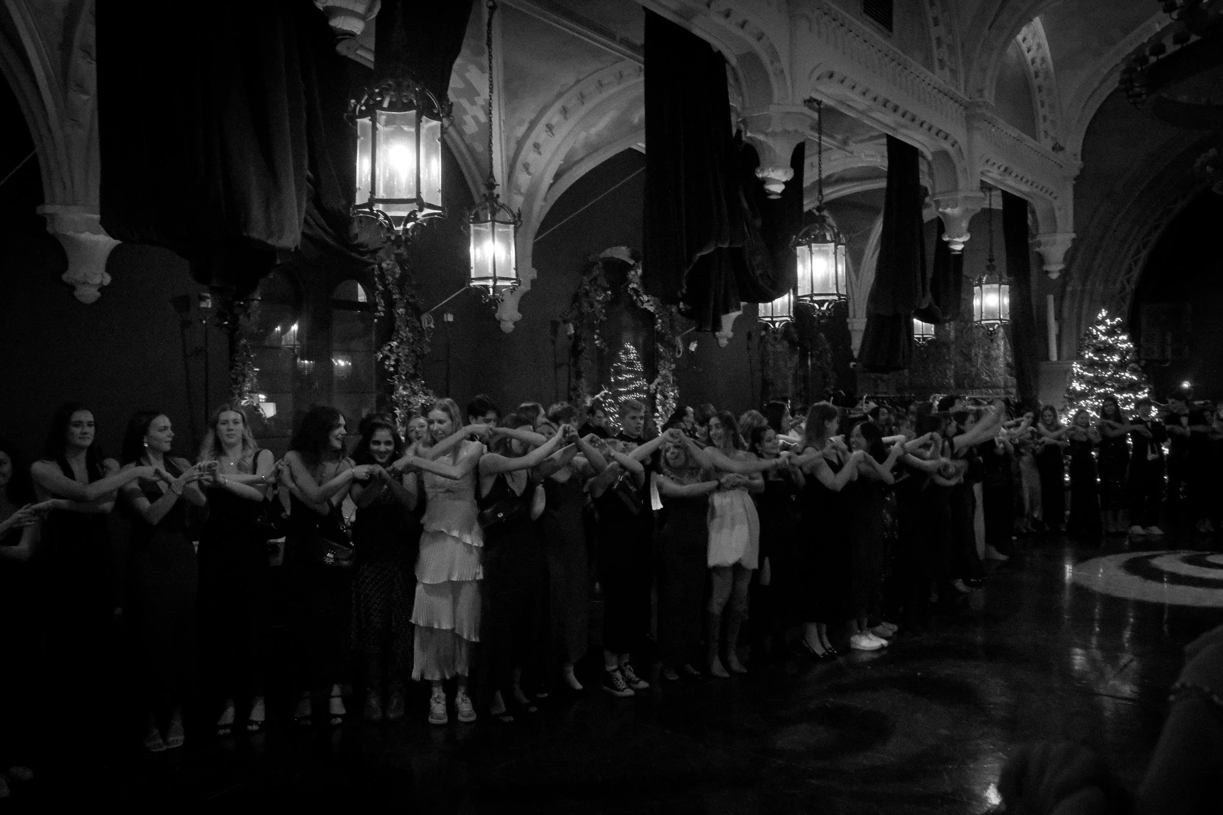 A group of people stand in a line holding hands inside a decorated hall with ornate architecture and Christmas trees.