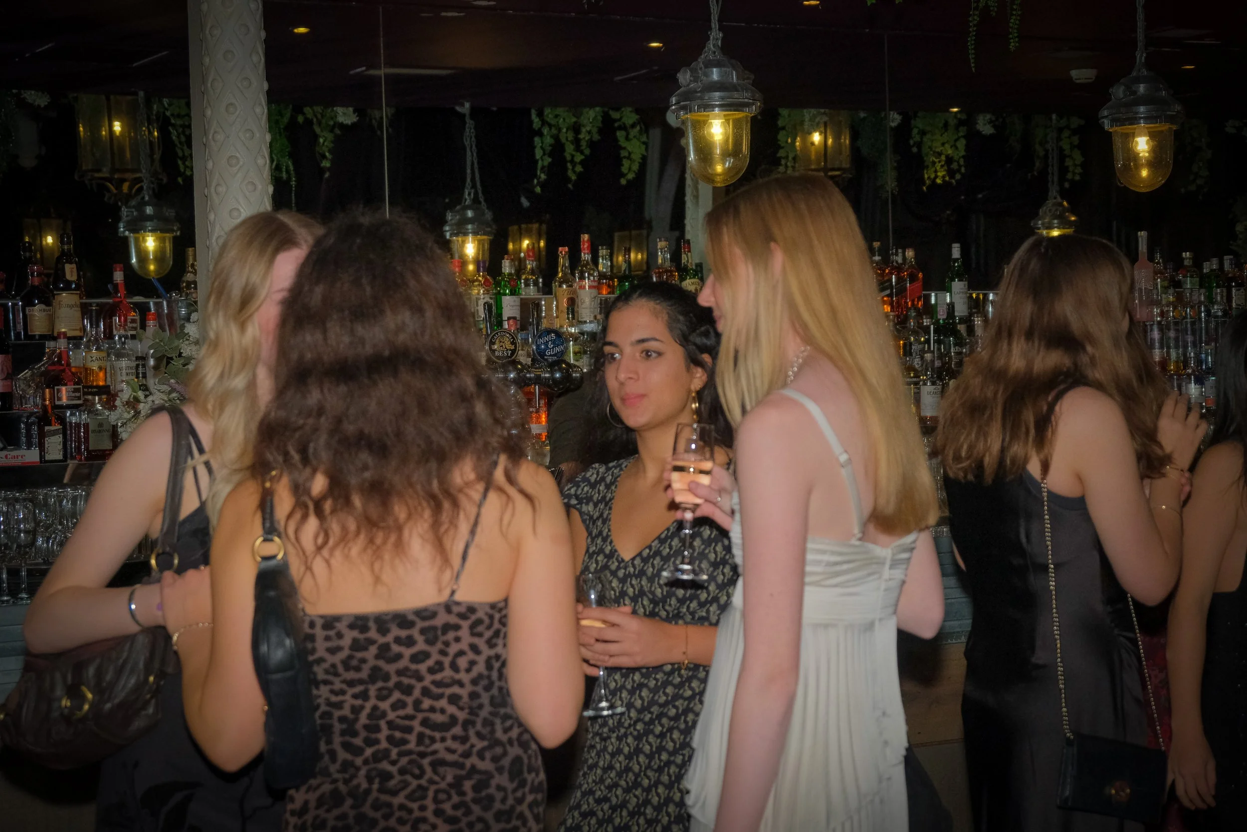 Group of women socializing at a bar, some holding drinks, in a dimly lit setting with bottles of alcohol in the background.