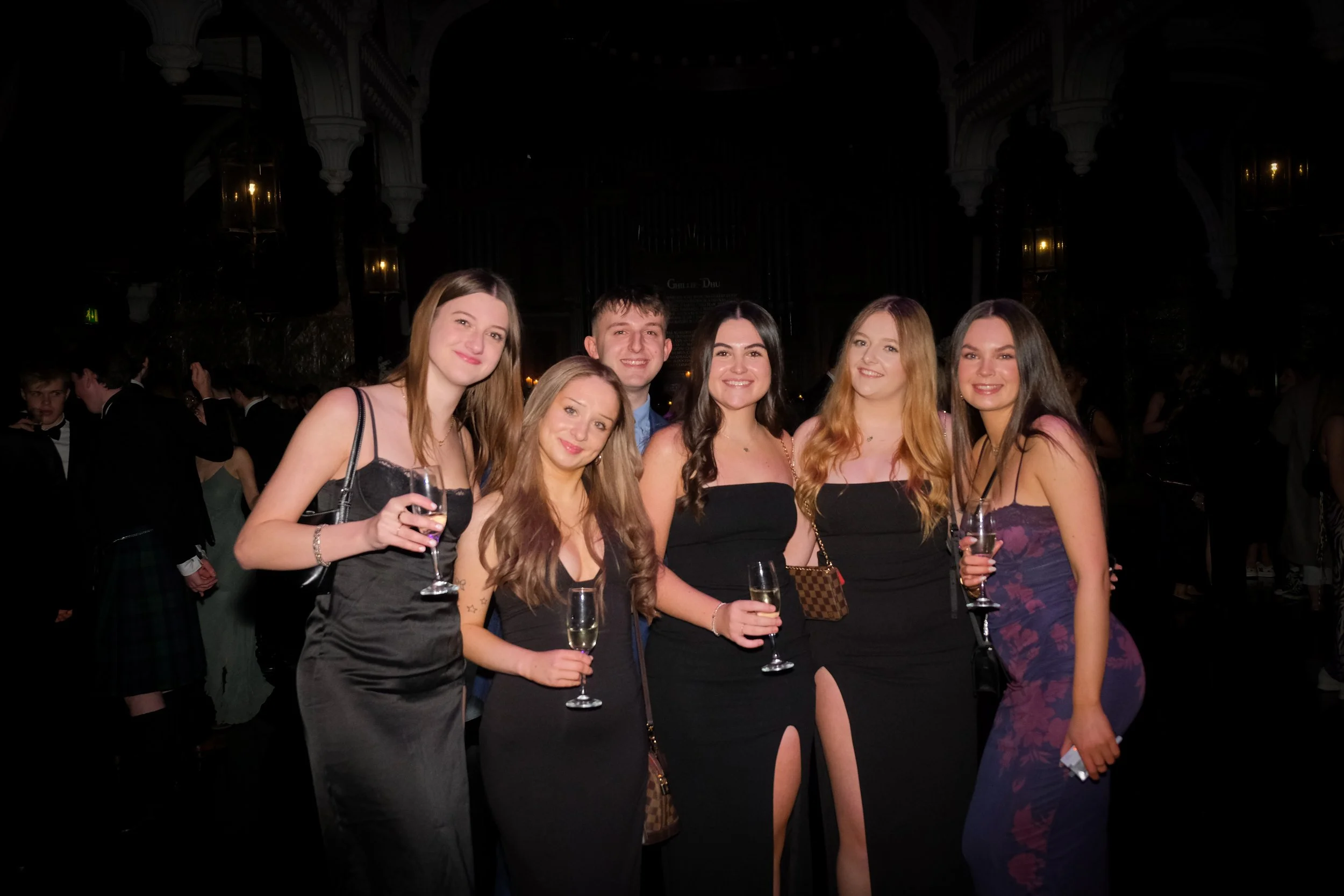 Group of six young adults dressed in formal evening wear at a social event, smiling and holding glasses of champagne, in an elegant indoors setting.