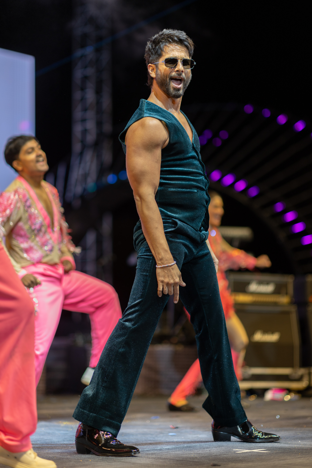 A man wearing sunglasses, a sleeveless velvet shirt, and black pants performing on stage with backup dancers in colorful outfits.
