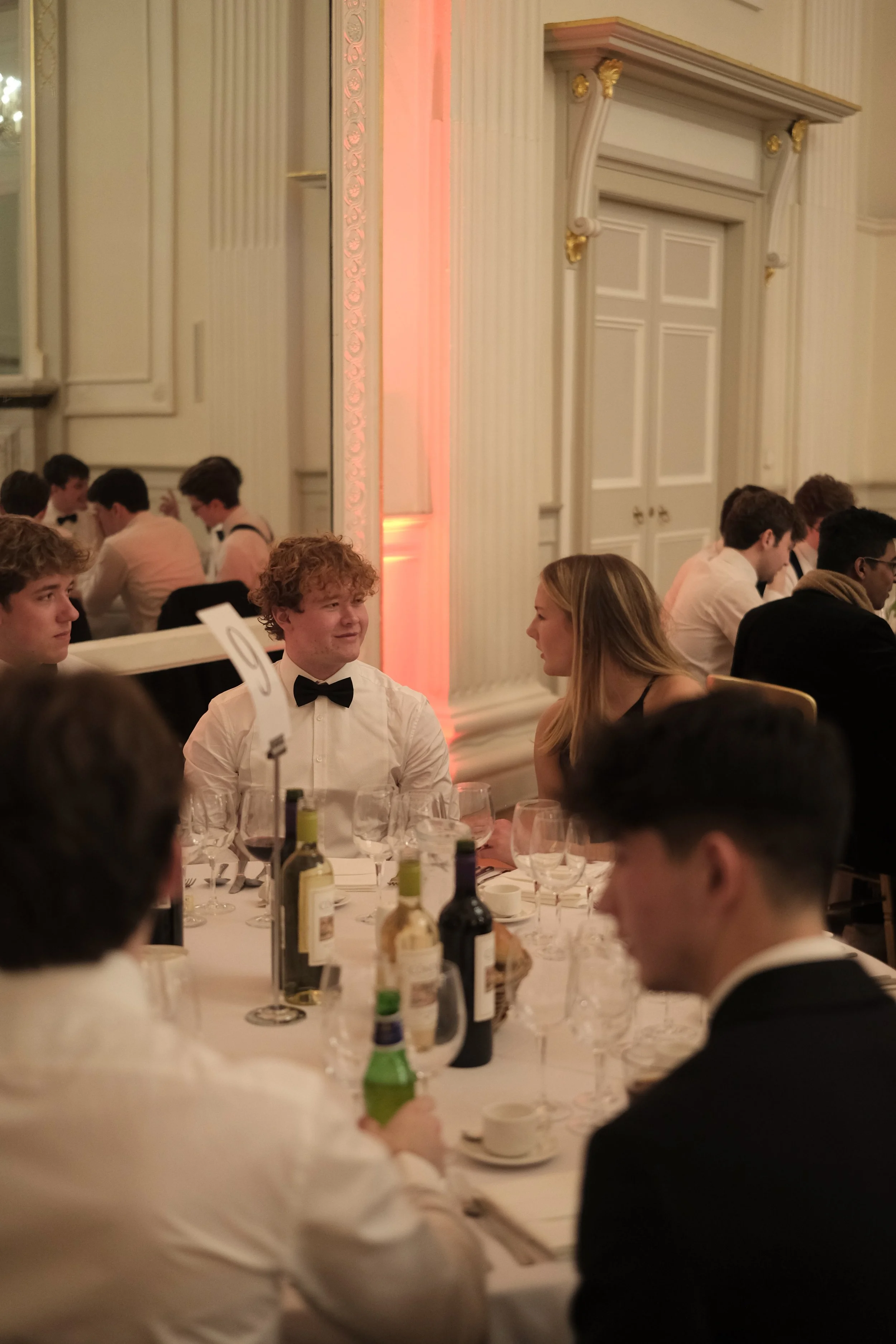 A group of young people dressed formally, sitting at a round banquet table with wine bottles, glasses, and tableware, in an elegant room with ornate details.