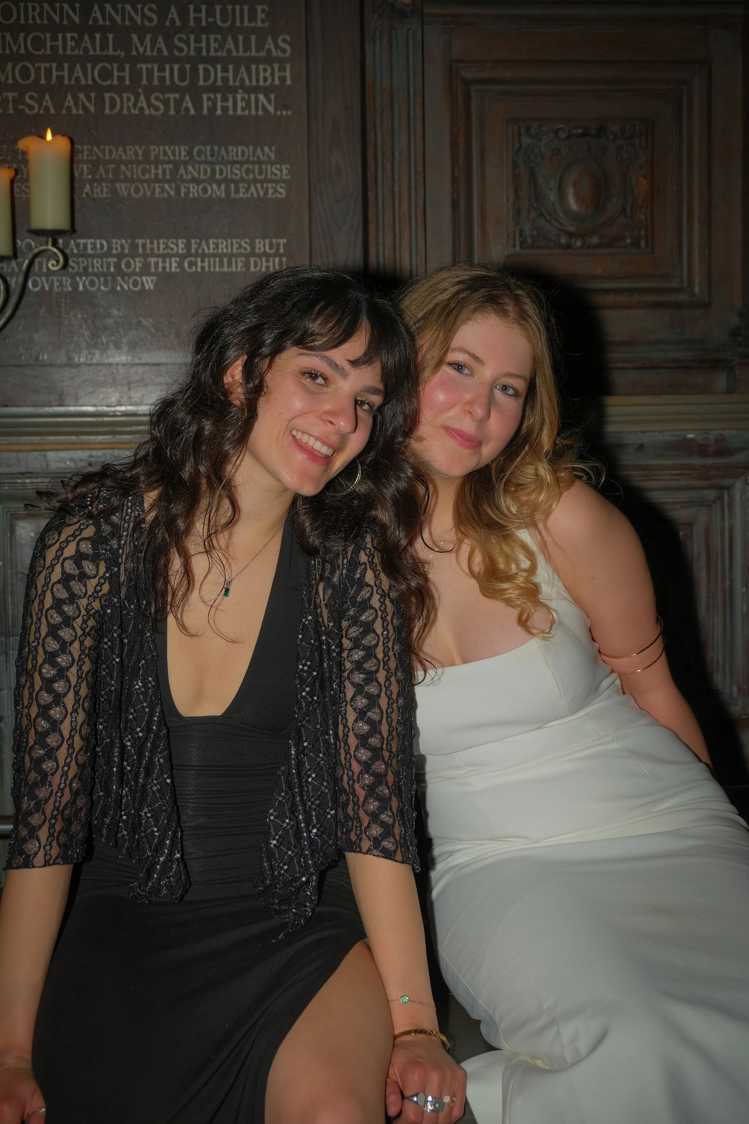 Two young women sitting close together in elegant dresses, smiling indoors with dark wooden paneling and a plaque with Gaelic text behind them.