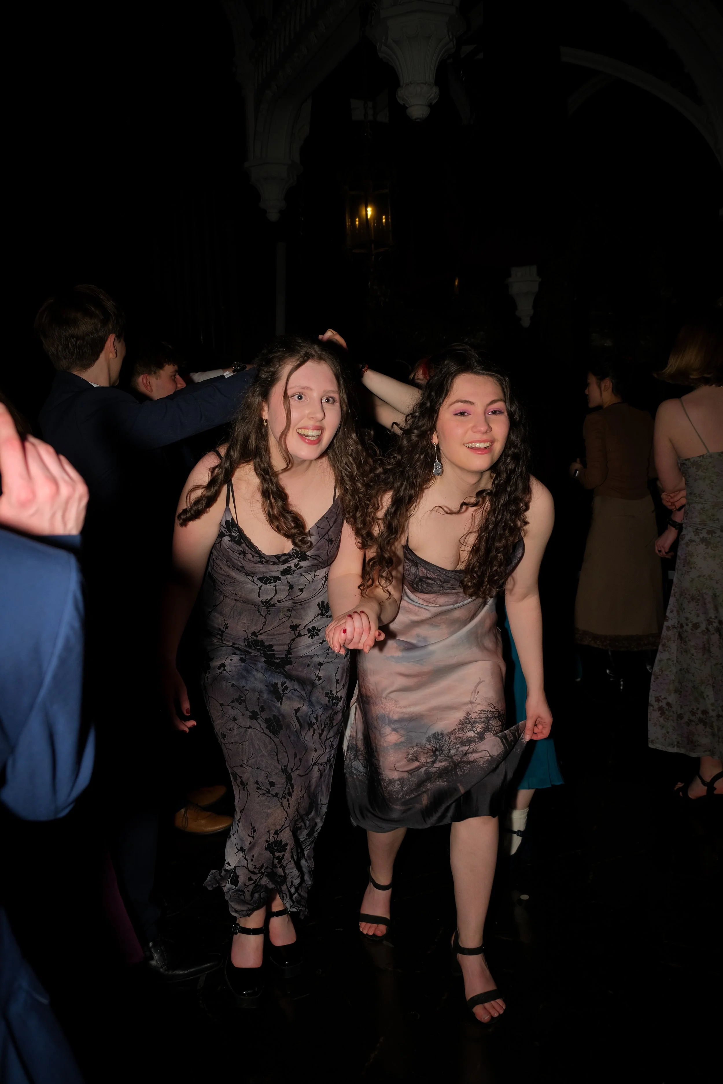 Two young women dancing and smiling at a party or celebration, surrounded by other guests in a dimly lit venue.