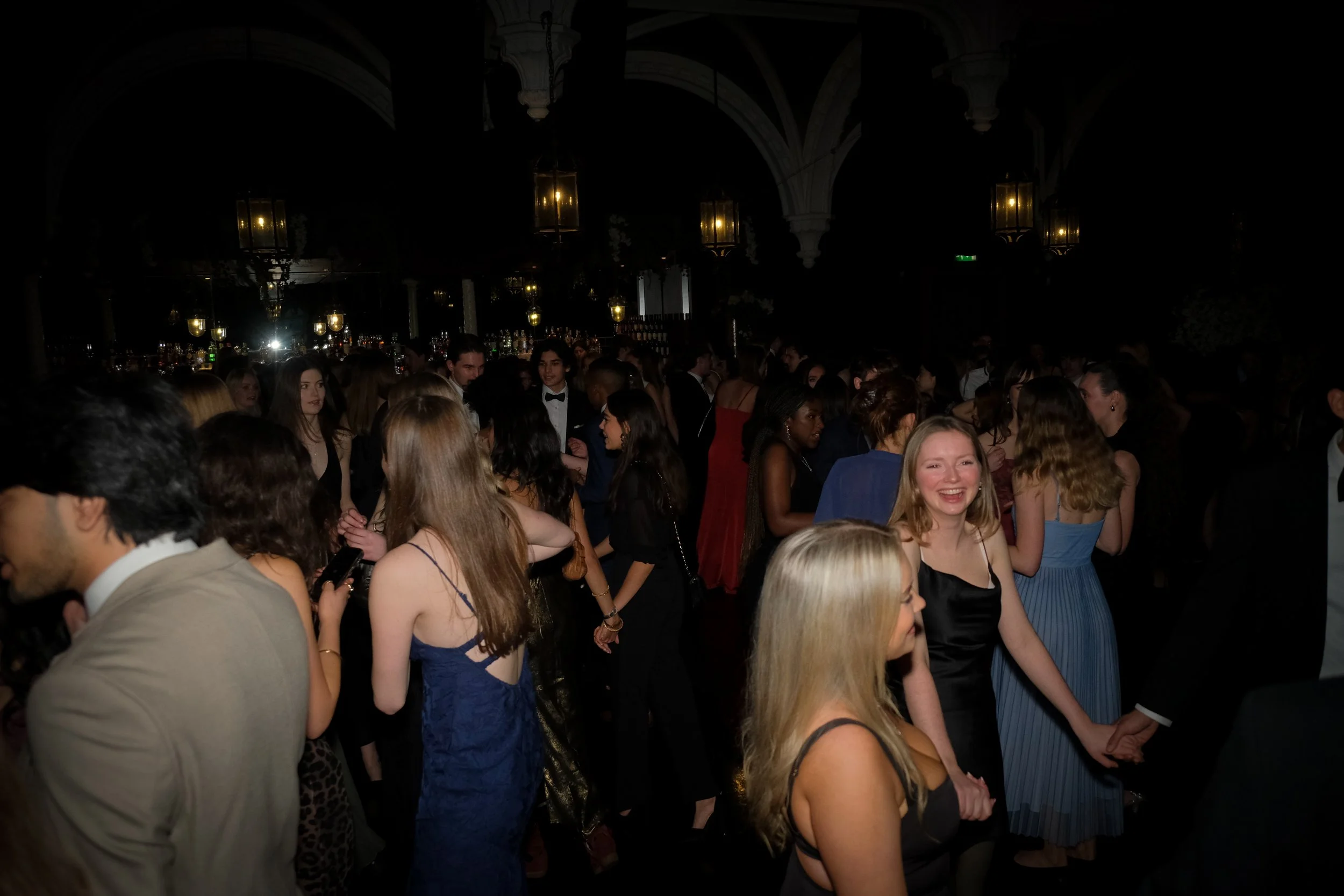 People dressed in formal attire at a party or dance event in a large, dimly-lit venue with chandeliers and bar area in the background.
