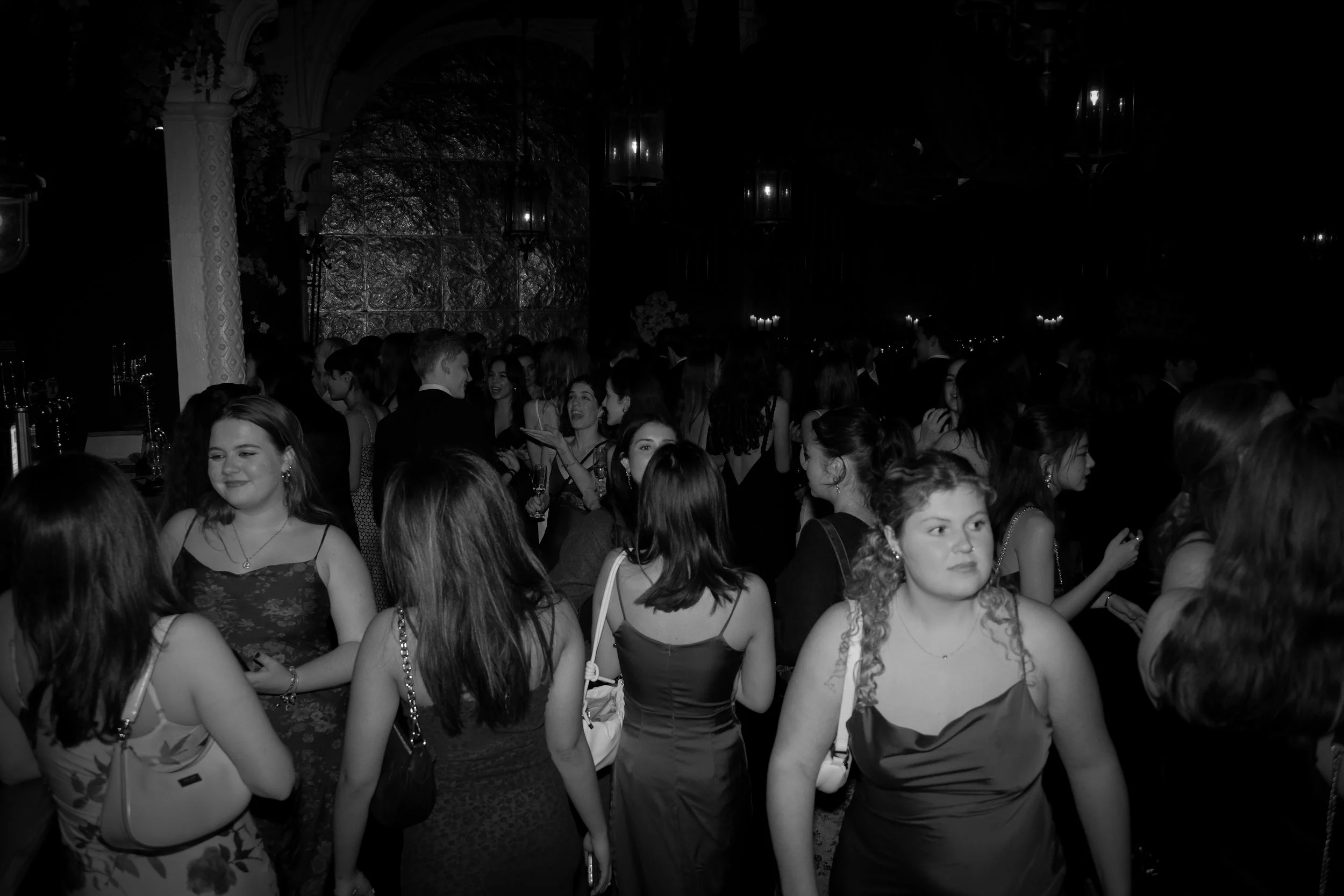 People gathered at a formal event or dance, dressed in elegant evening attire, in a dimly lit setting with ornate decor.
