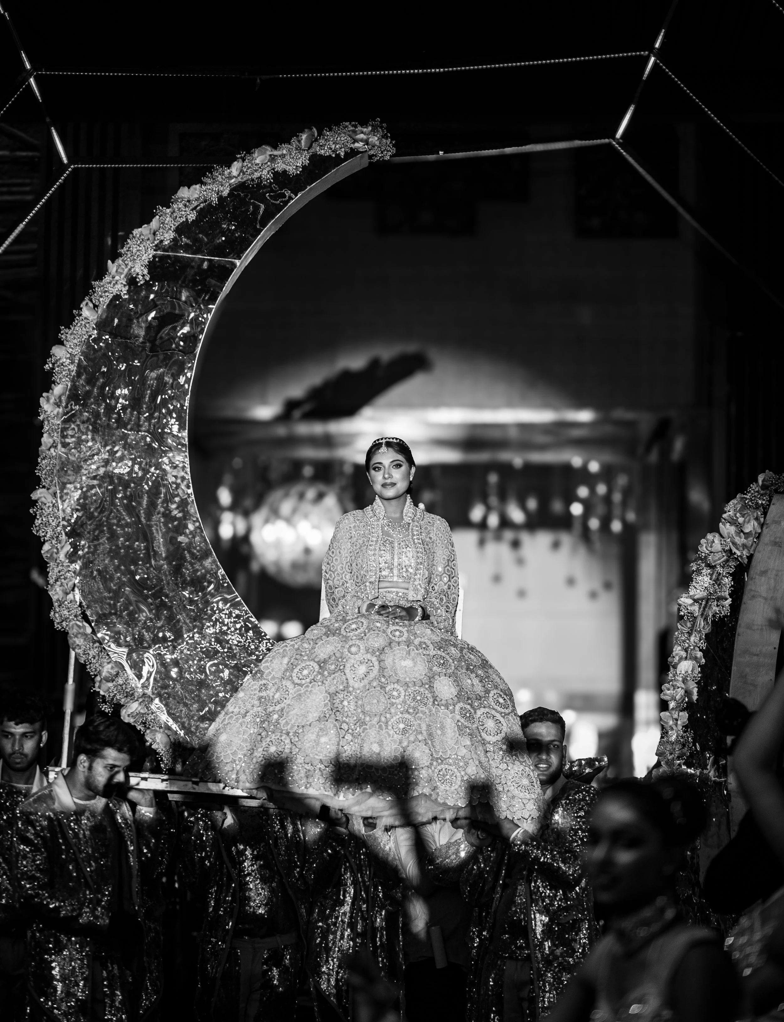 A woman dressed in traditional Indian attire sitting on a decorated float shaped like a crescent moon during a festival or parade, surrounded by people.