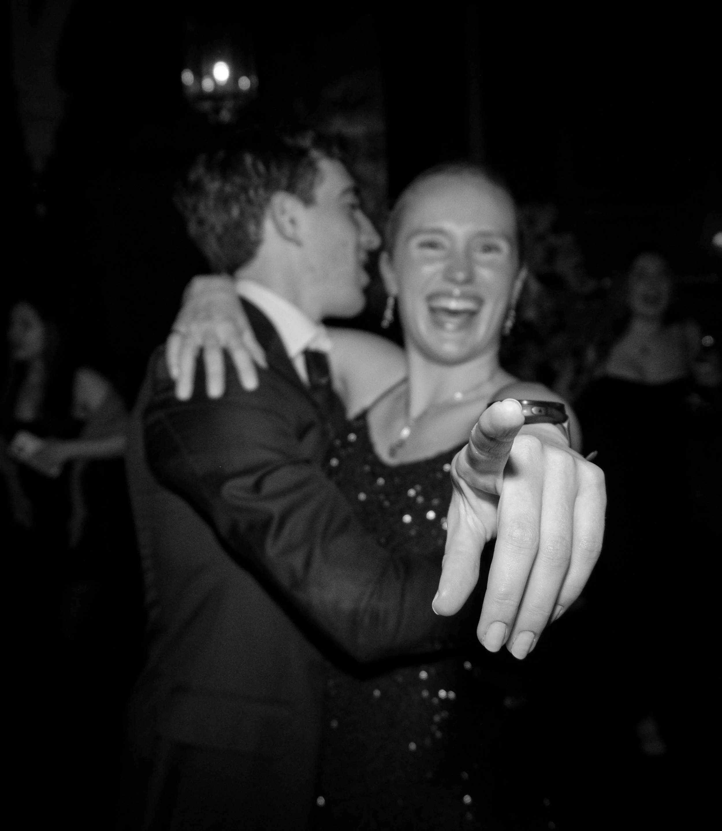 A black and white photo of a dancing couple at a formal event. The woman is smiling and holding a phone, while the man is kissing her cheek.