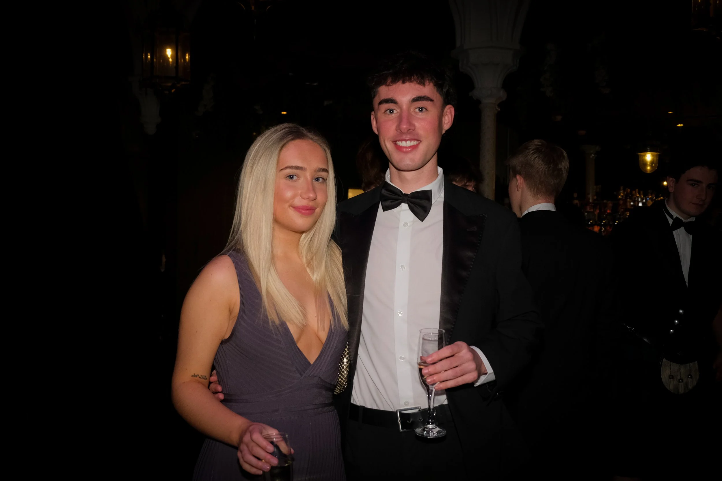 A young man and woman dressed in formal attire at a social event, holding glasses of drinks and smiling at the camera.