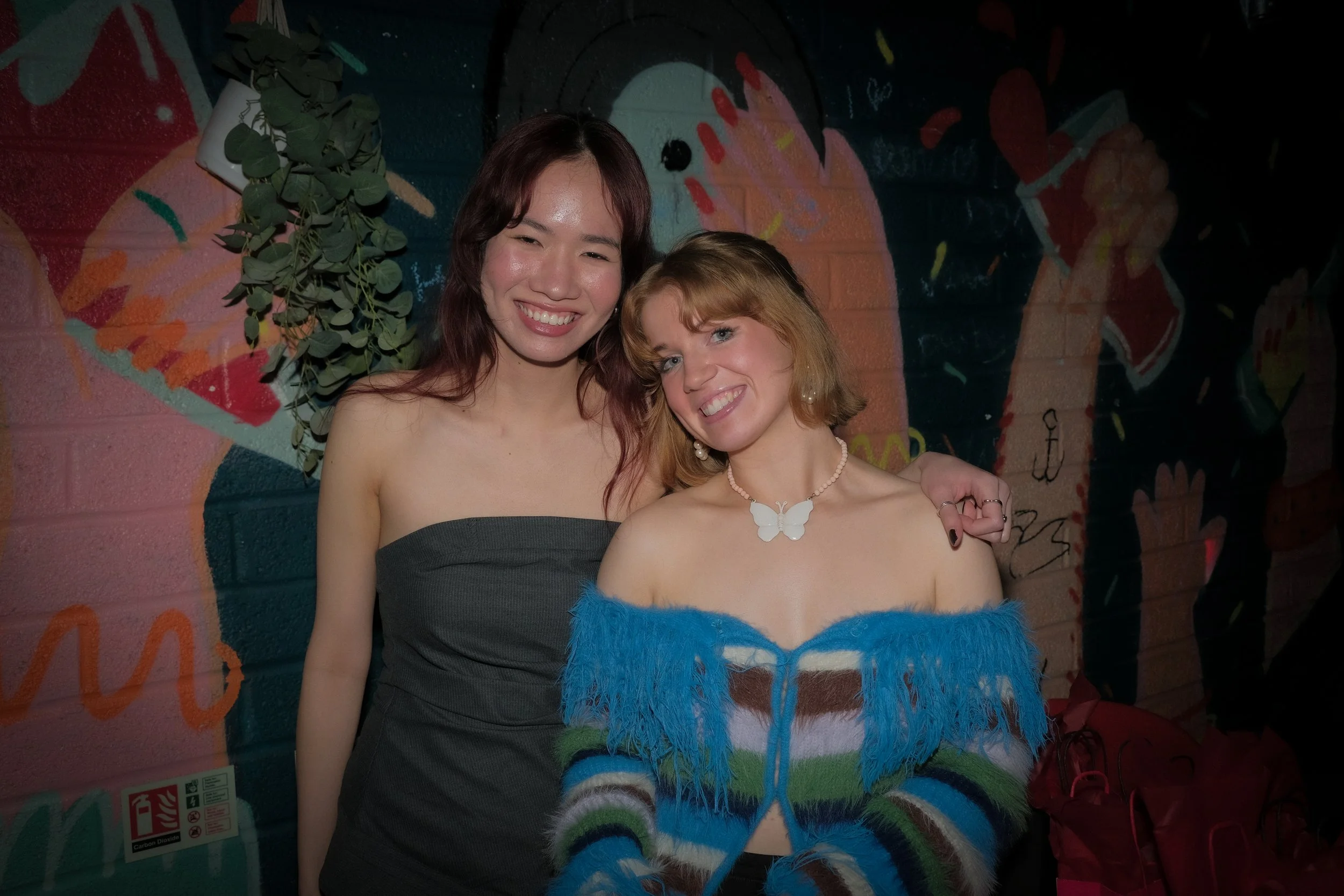 Two women smiling and standing close together in front of a colorful, graffiti-style mural on a brick wall. One woman has long dark hair and is wearing a strapless black dress, while the other has short, wavy red hair and is wearing a blue, off-the-s