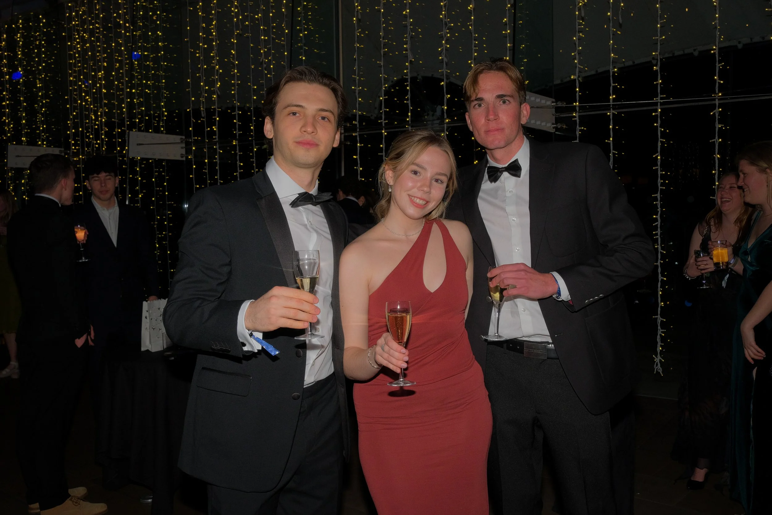 Three young adults in formal attire at a celebration, holding glasses of champagne, smiling for the photo, with decorative string lights in the background.