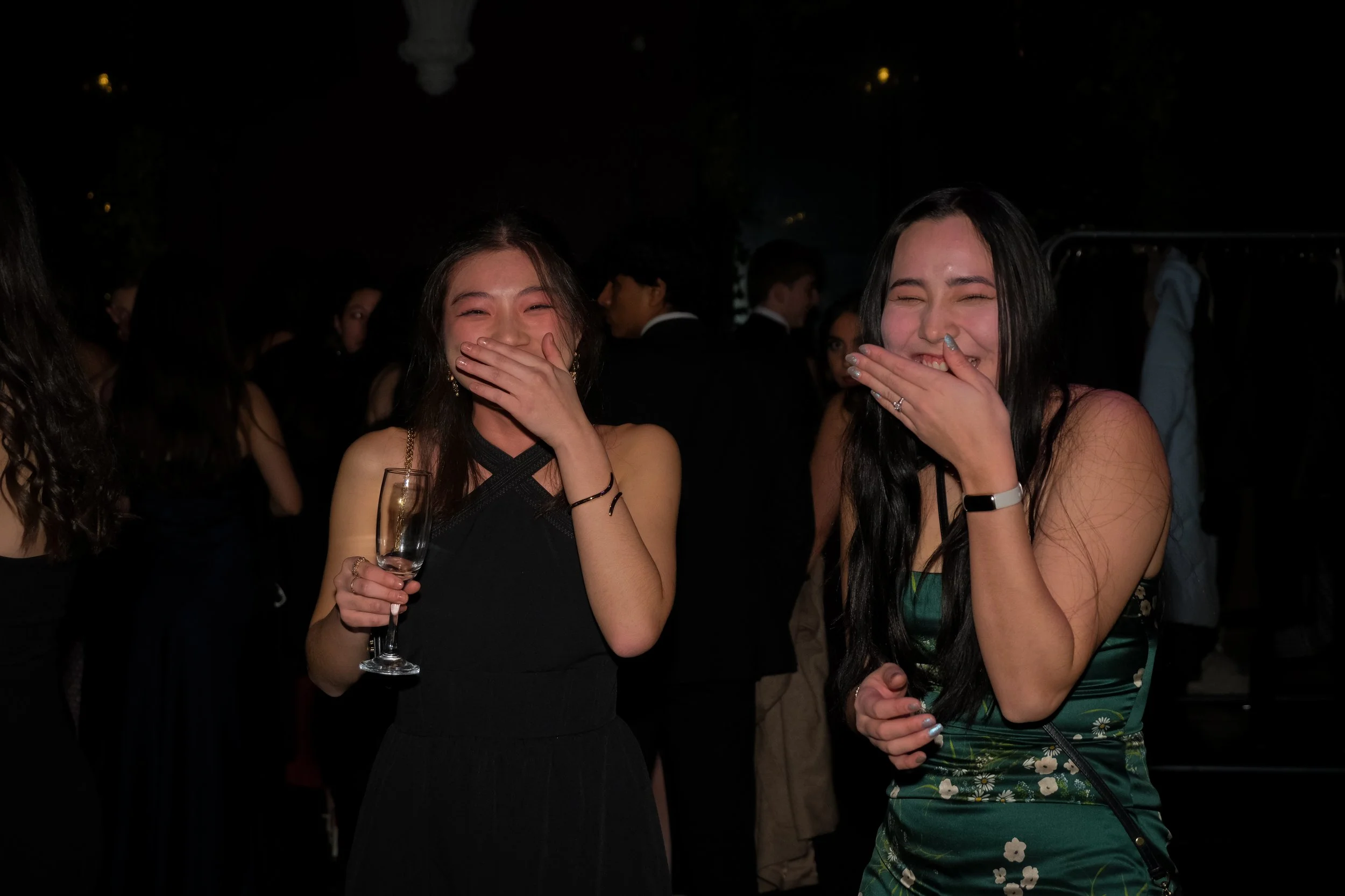 Two women in formal attire laughing and covering their mouths in a dark setting, with other people in the background.