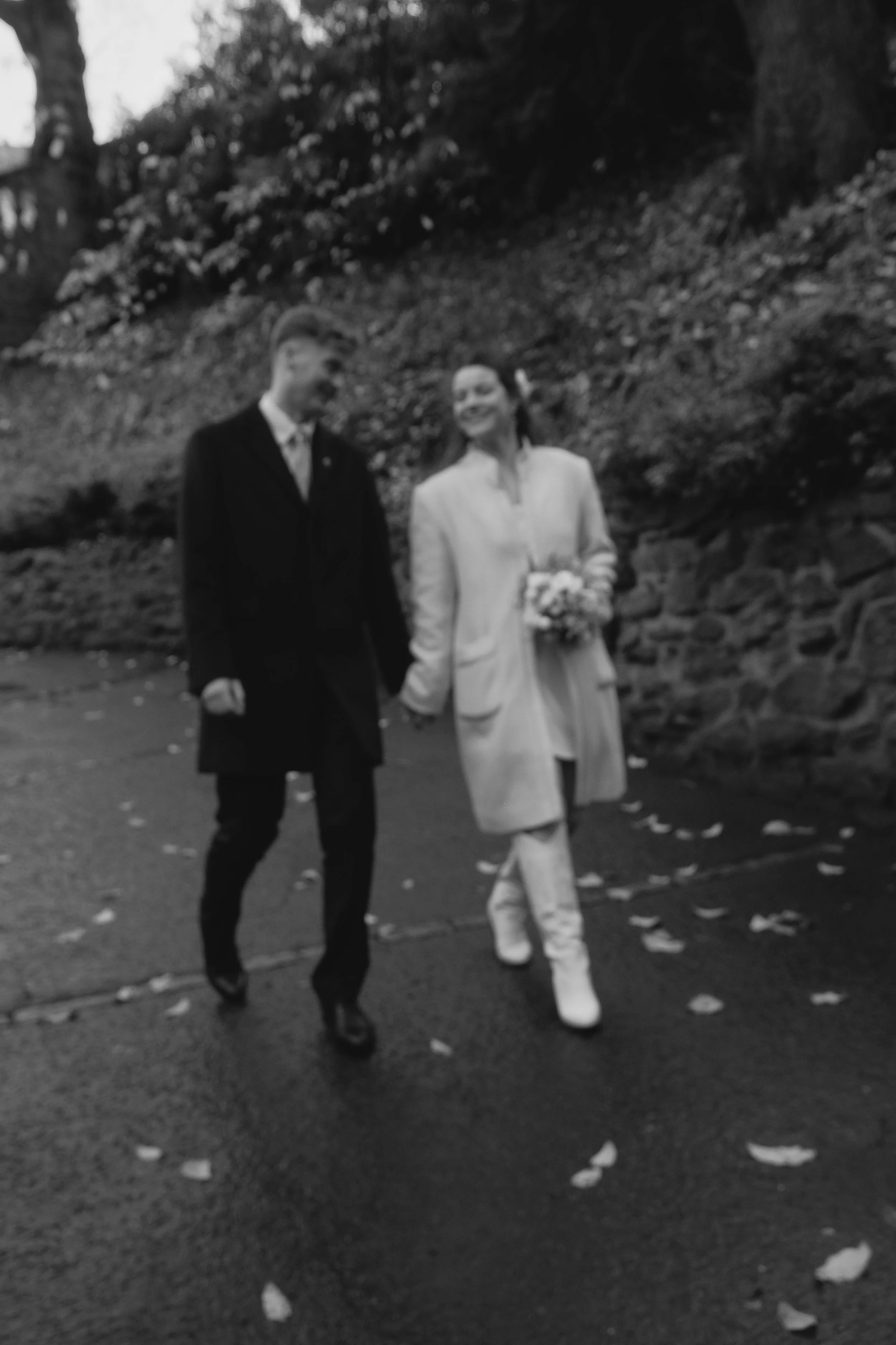 A black and white photo of a couple walking outdoors, holding hands. The woman is dressed in a light coat and boots, and is holding a bouquet of flowers. The man is in a dark formal suit. Leaves are scattered on the path, and there is a stone wall an