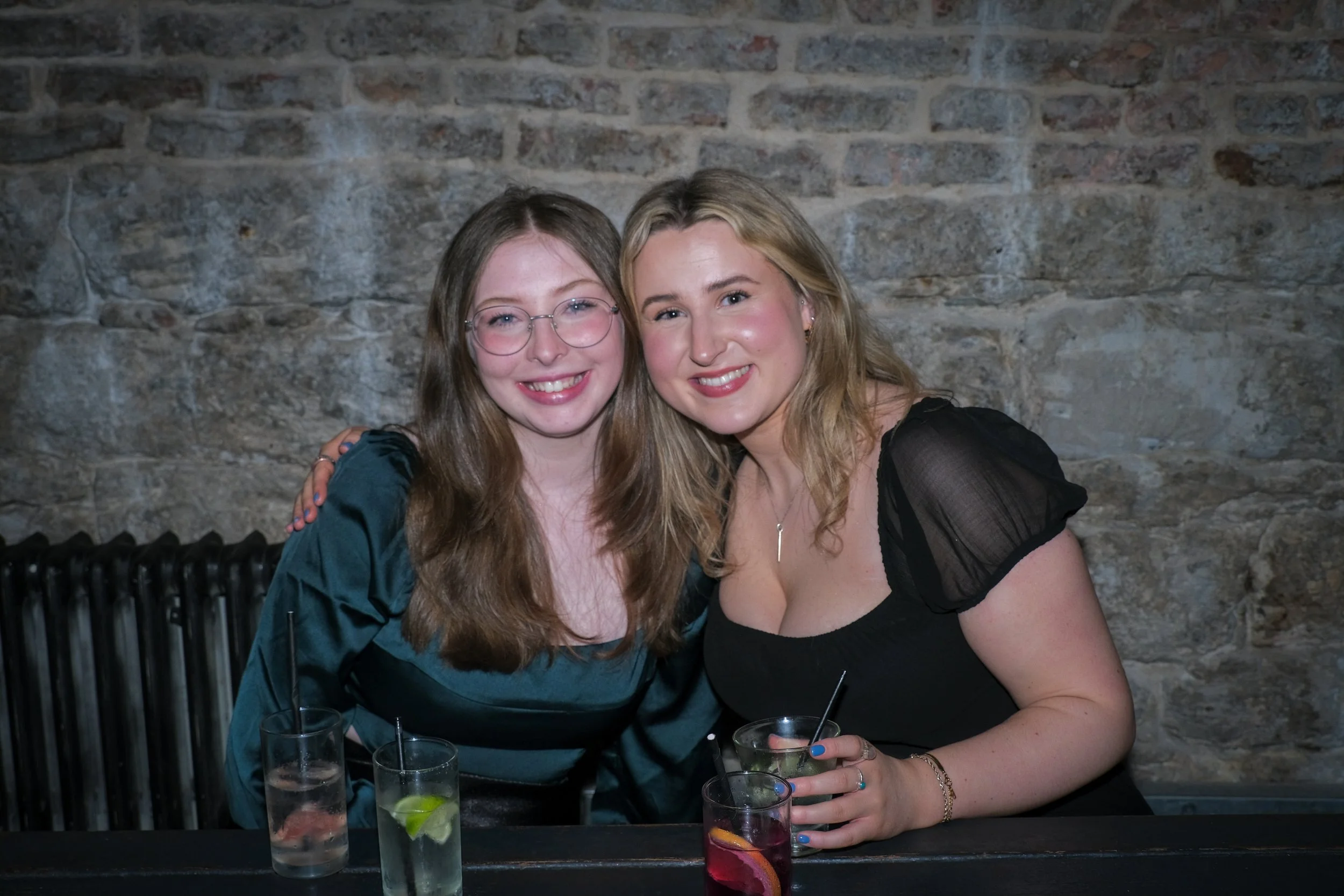 Two smiling women sitting together at a bar with drinks, against a brick wall background.