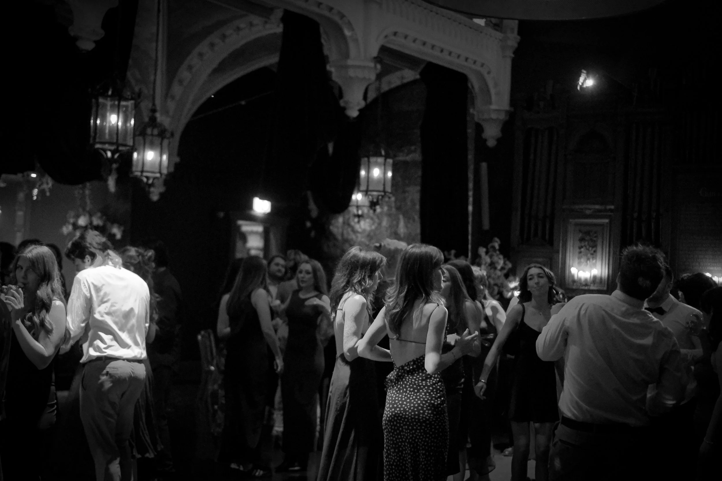People dancing and socializing at a formal event or party in a dimly lit venue with ornate decorations and wall sconces.