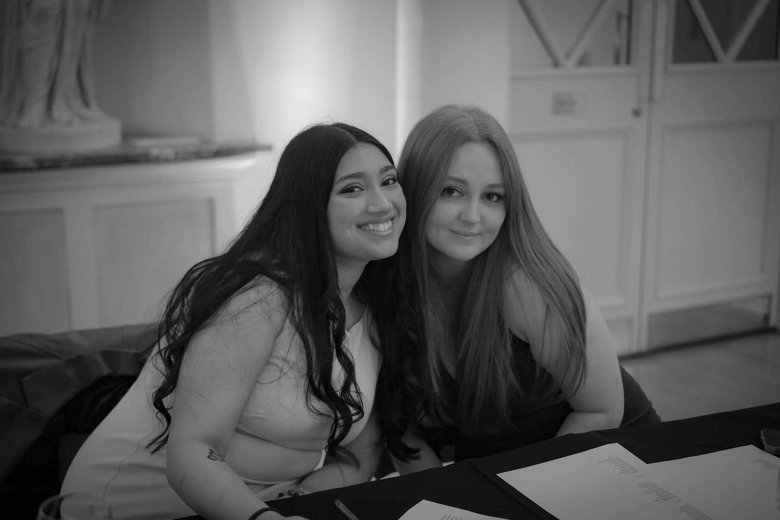 Black and white photo of two smiling women sitting at a table, with papers in front of them, in an indoor setting.