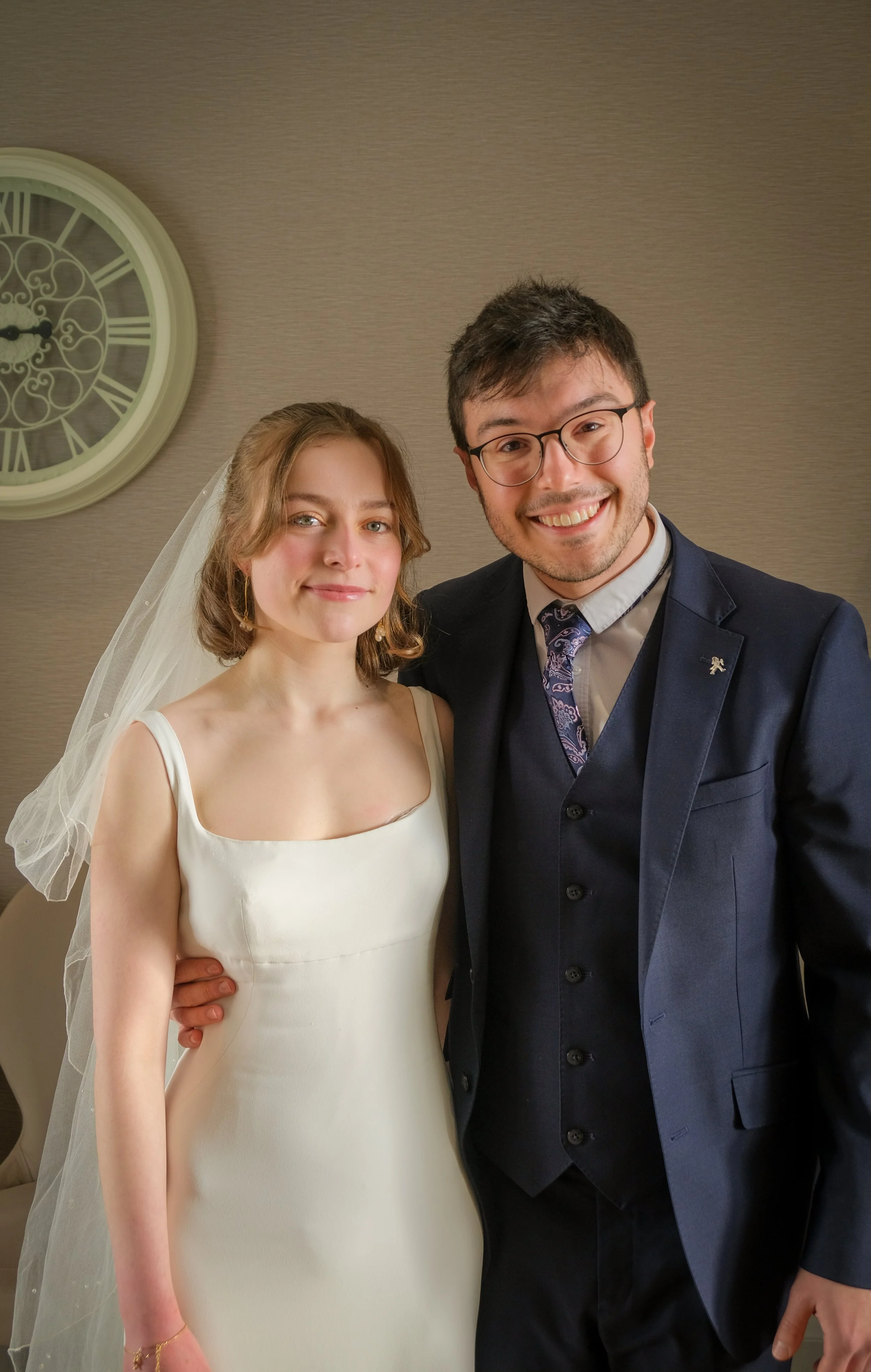 A bride and groom standing together indoors, smiling at the camera. The bride is wearing a white wedding dress with a veil, and the groom is dressed in a navy blue suit with a tie and glasses. A large clock is visible on the wall behind them.