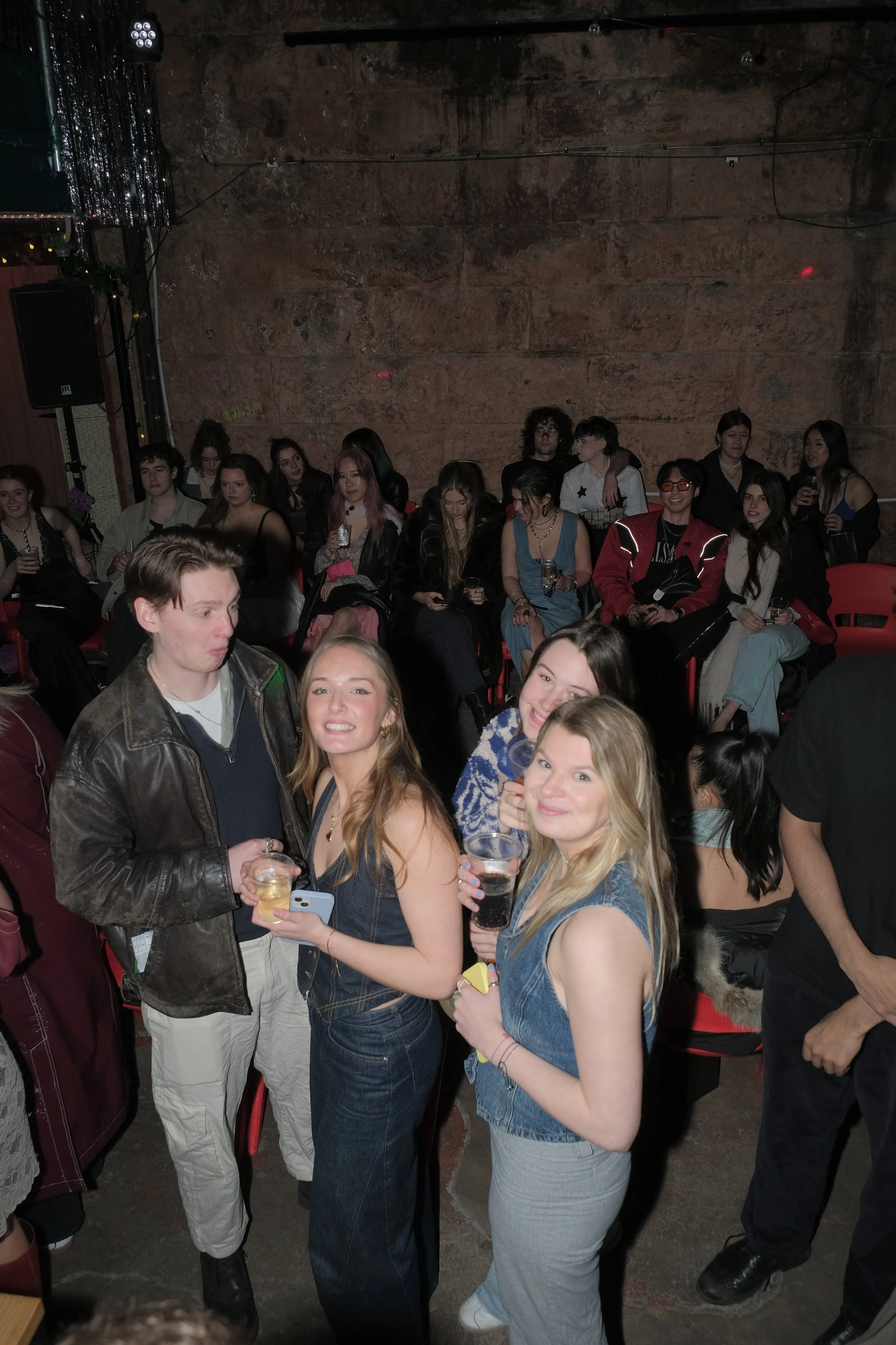 People at a party, some holding drinks, with a group sitting in the background in a dimly lit venue.