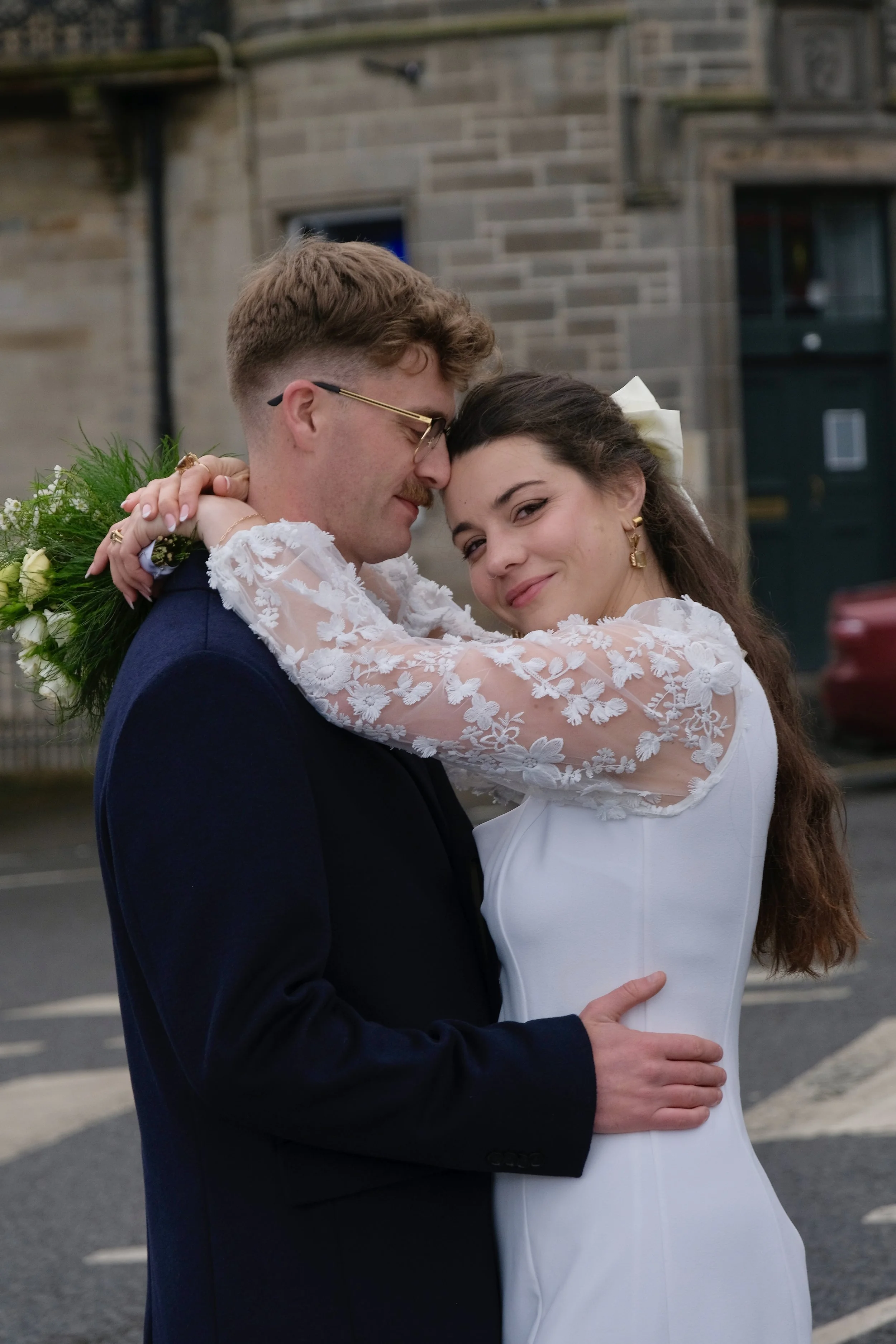 A newlywed couple embracing on a city street with stone buildings in the background. The groom is in a dark suit, and the bride is in a white dress with lace sleeves.
