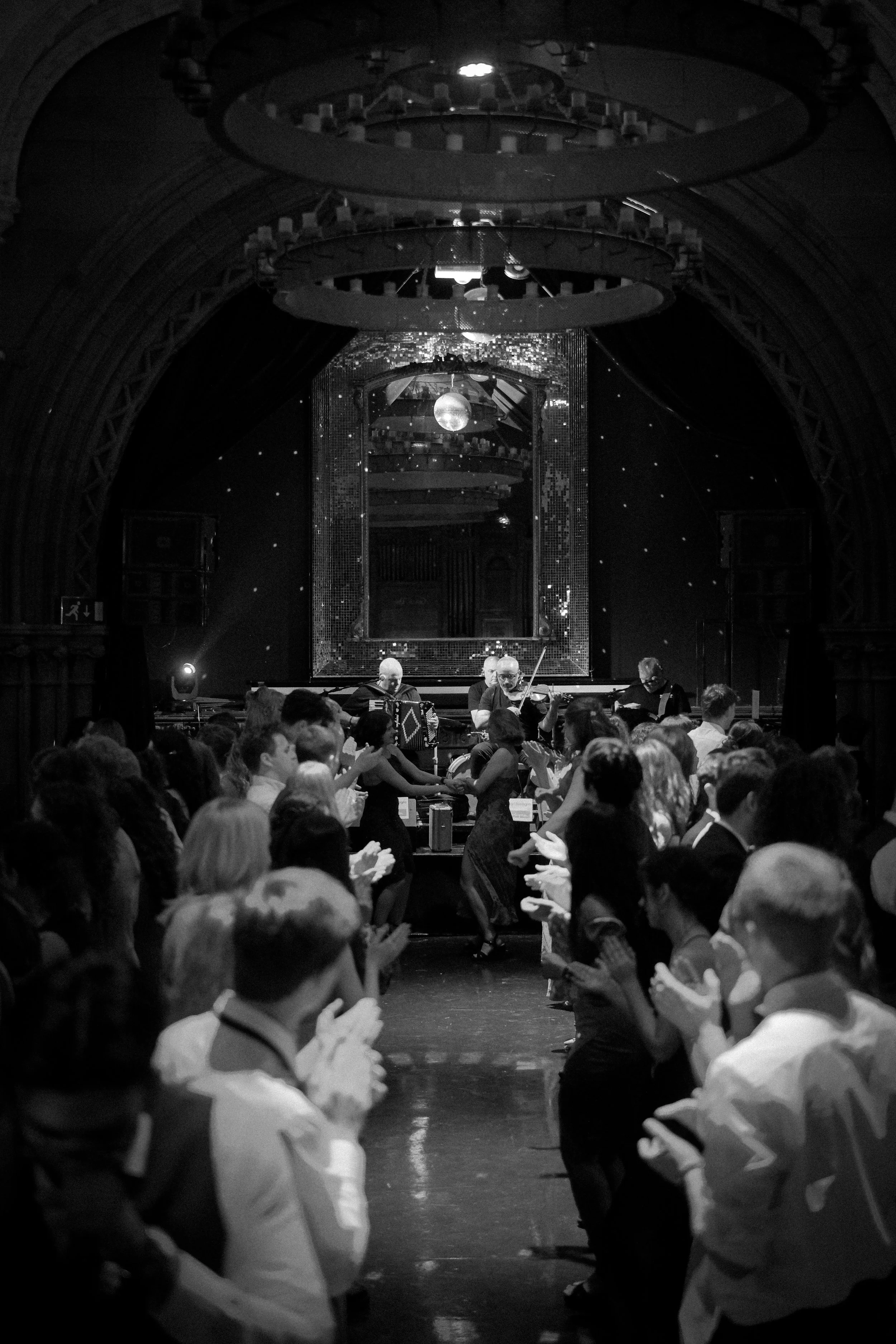 People dancing in a ballroom with a live band playing on stage, decorated with mirrors and starry background.