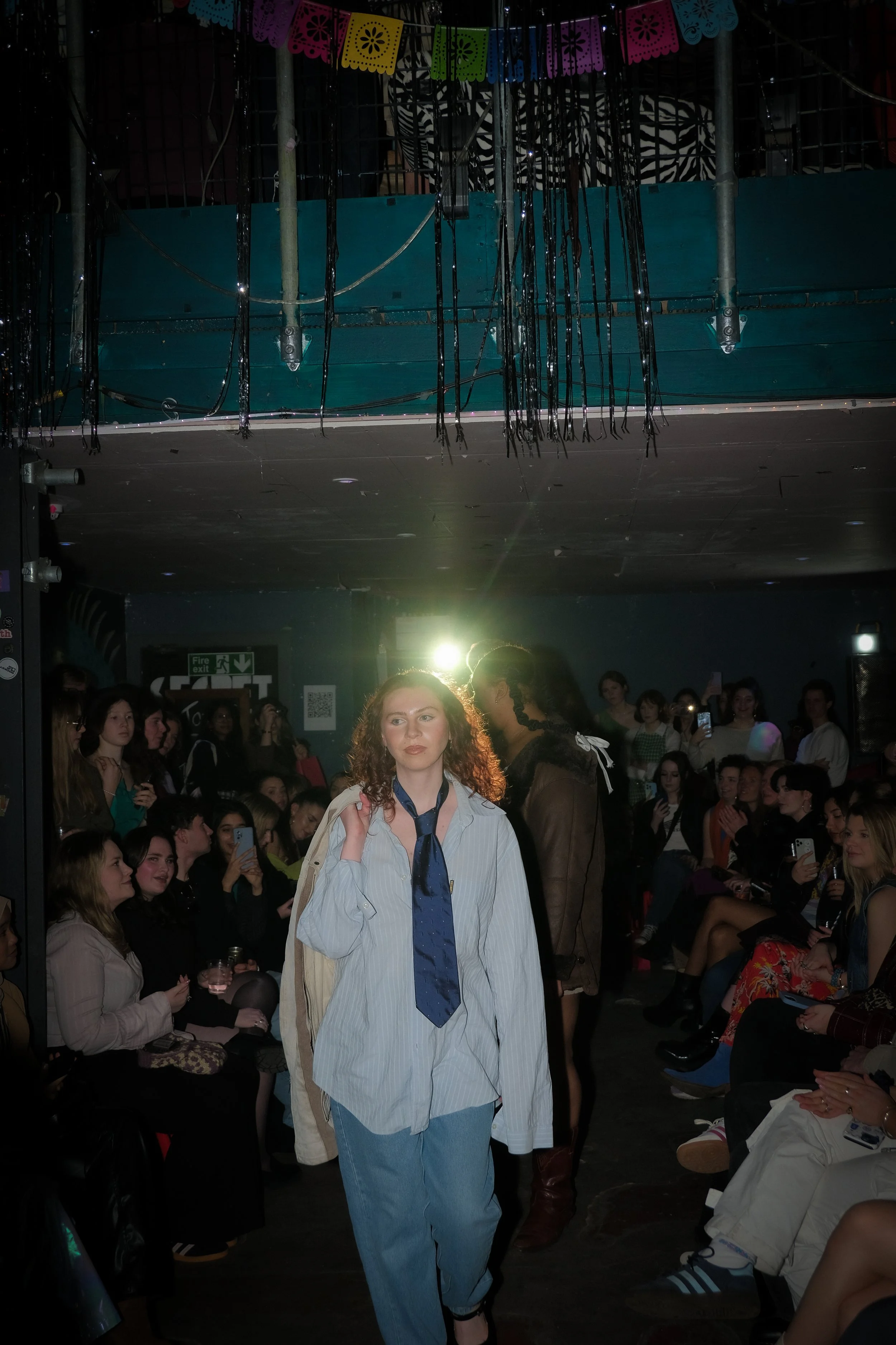 A woman with curly hair walking on a runway during a fashion show. She is wearing a loose shirt with a tie, and the audience is seated on both sides, taking pictures and watching.