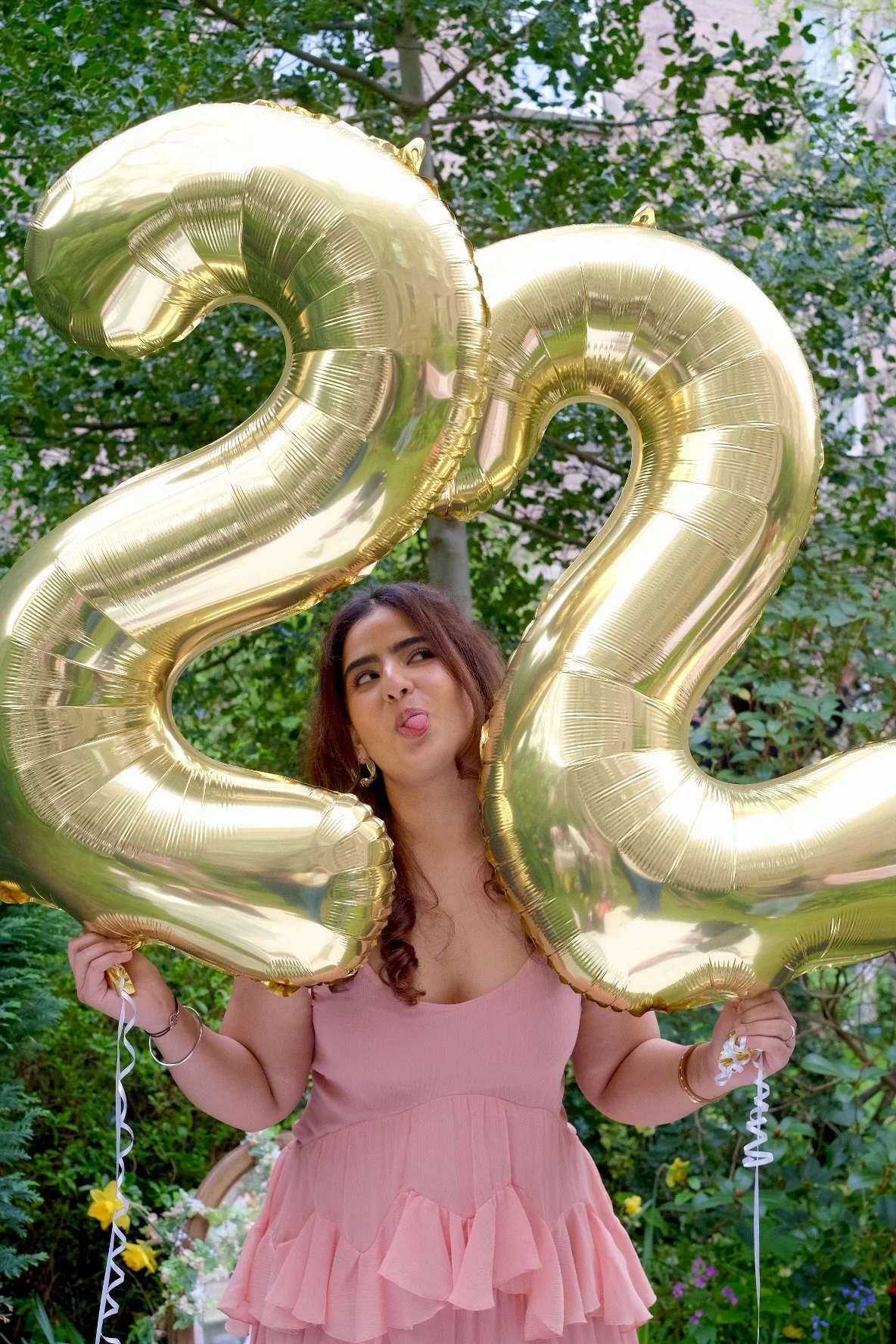A young woman in a pink dress holding large gold '2022' balloons outdoors, making a silly face with her tongue sticking out, behind green trees.