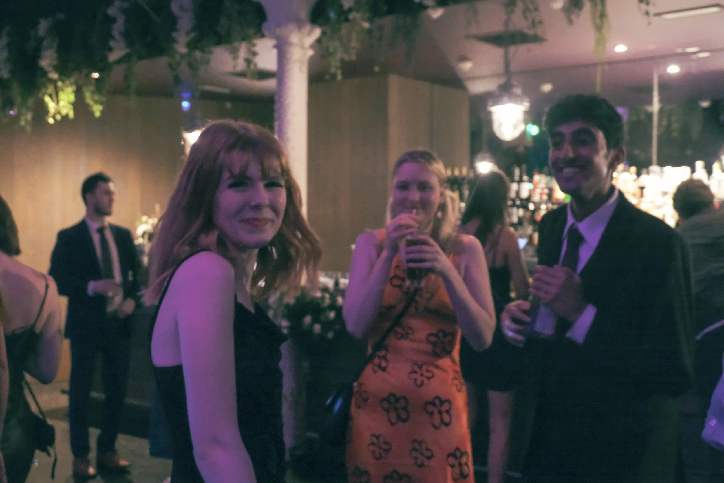 People at a social event, three are clearly visible in the foreground: a woman with red hair in a black dress, a woman in a sleeveless orange dress with black floral patterns holding a drink, and a man in a suit with a tie, smiling at the camera. Bac