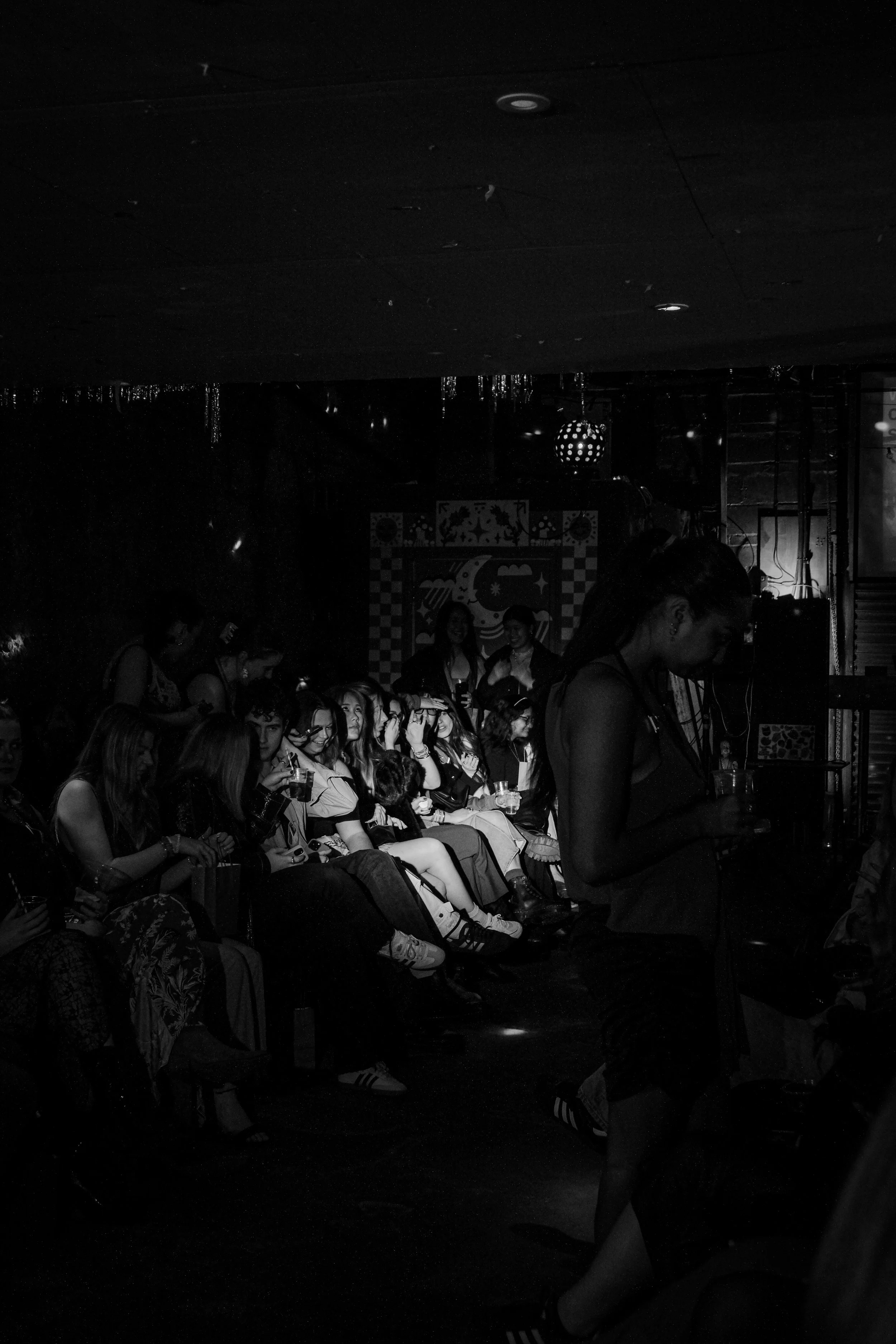 A group of young women sitting closely together in a dark room, some wearing glasses, with a woman standing in the foreground. The scene appears to be a social gathering, possibly at a nightclub or party, with dim lighting and decorative background.