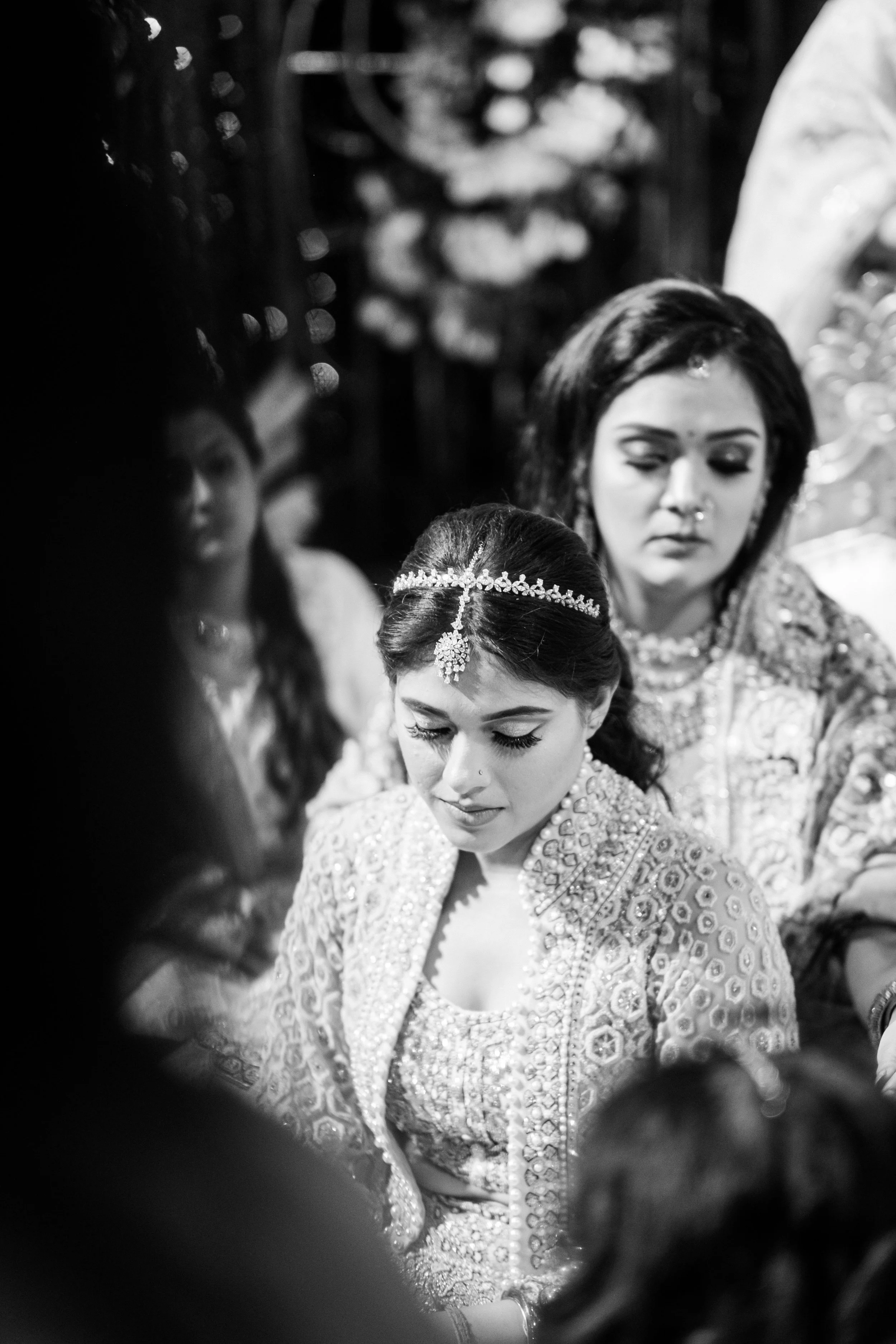 A black and white photograph captures a bride and another woman, likely a bridesmaid, participating in a traditional ceremony. The bride is wearing intricate jewelry and a heavily embroidered dress, with her head bowed. The woman next to her is also