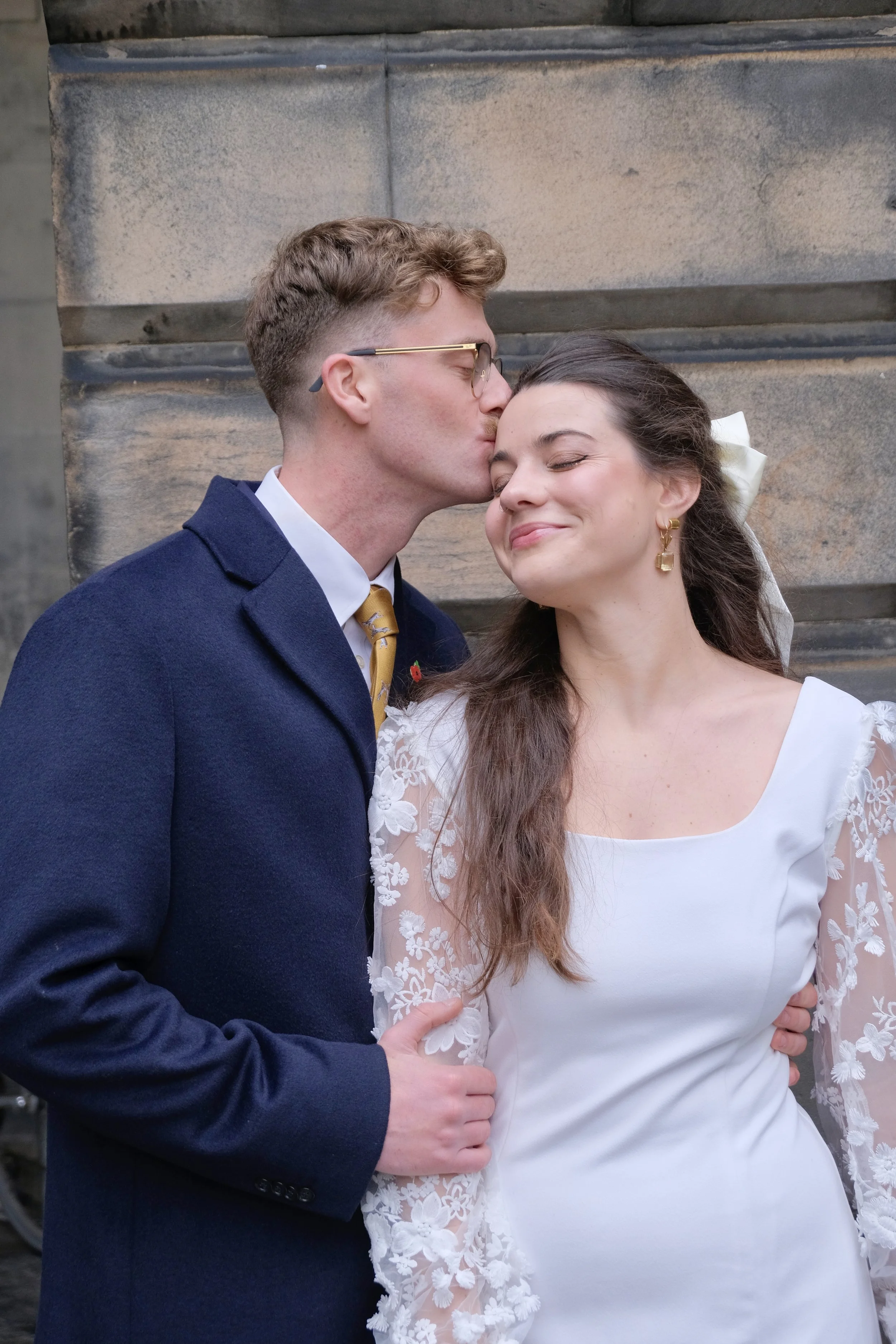 A man in a navy coat and yellow tie kisses a woman in a white dress with lace sleeves on the forehead during a wedding or special occasion, standing in front of a stone wall.