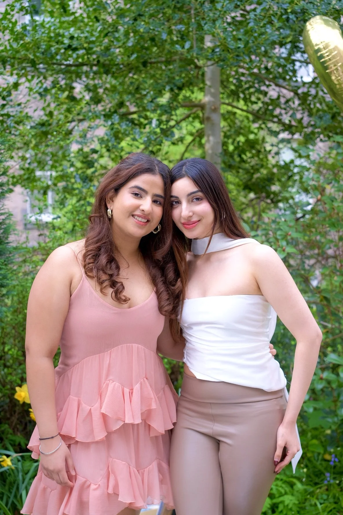 Two women standing close together outdoors, smiling, with green foliage in the background. One woman is wearing a pink ruffled dress, and the other is wearing a white top with beige pants.