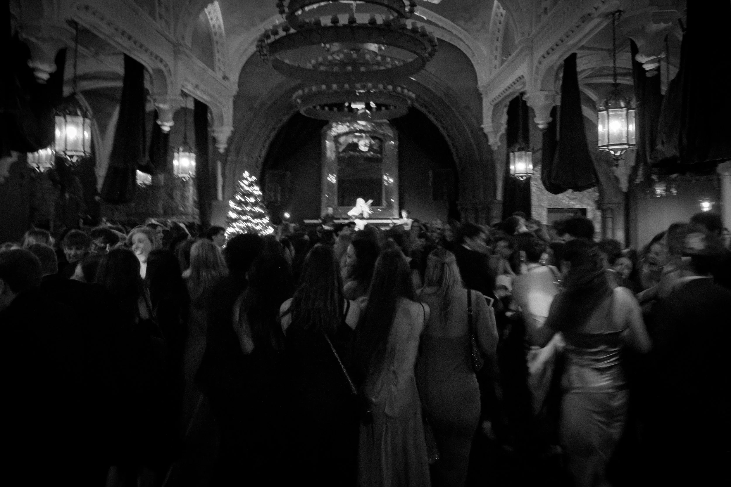 A crowded theater or banquet hall decorated for Christmas, with a Christmas tree, ornate architecture, chandeliers, curtains, and a stage with a performer playing a keyboard.