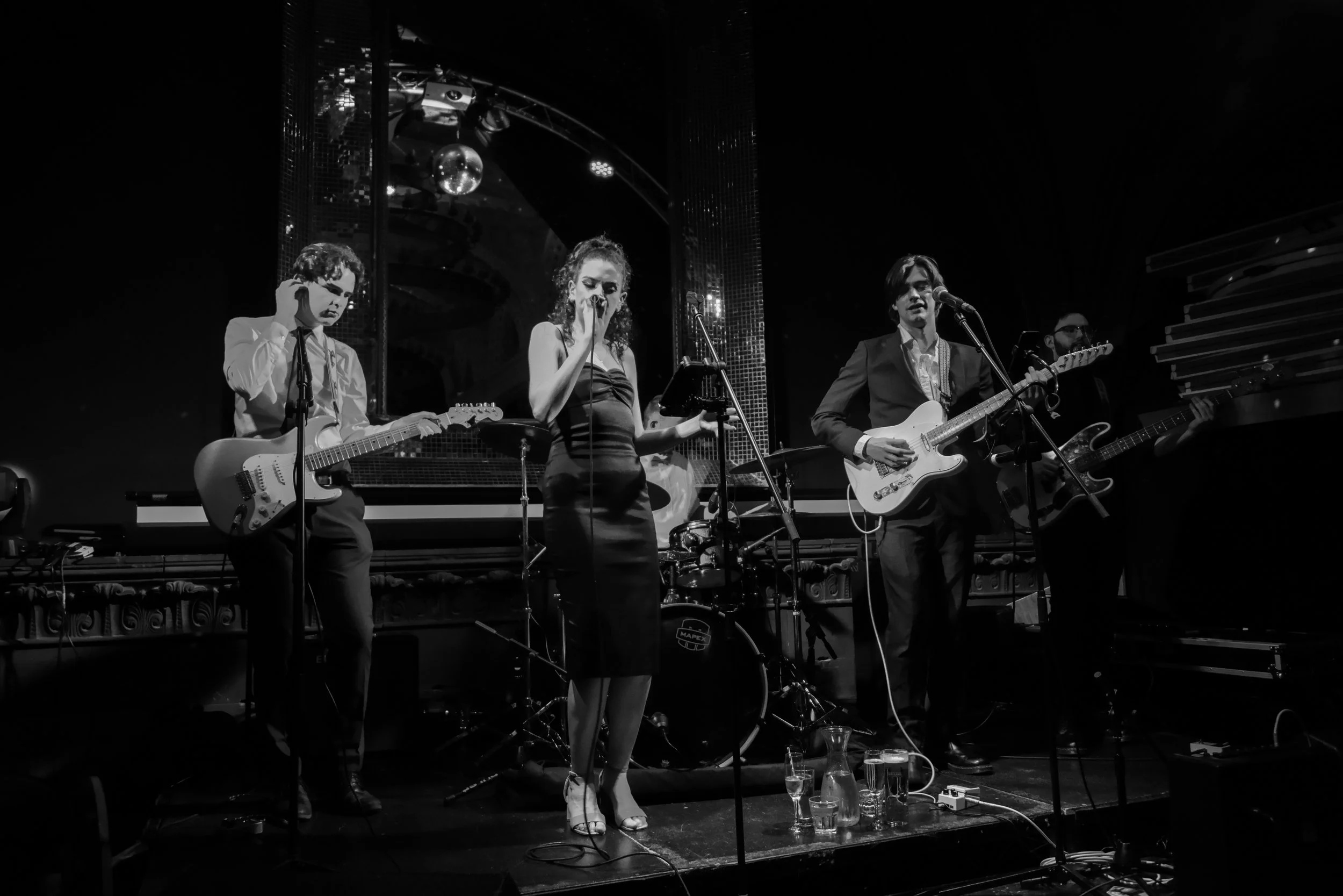 A black and white photo of a live band performing on stage with four musicians: a guitarist on the left, a female singer in the center, a guitarist on the right, and a bassist partially visible in the background. The stage has microphones and musical