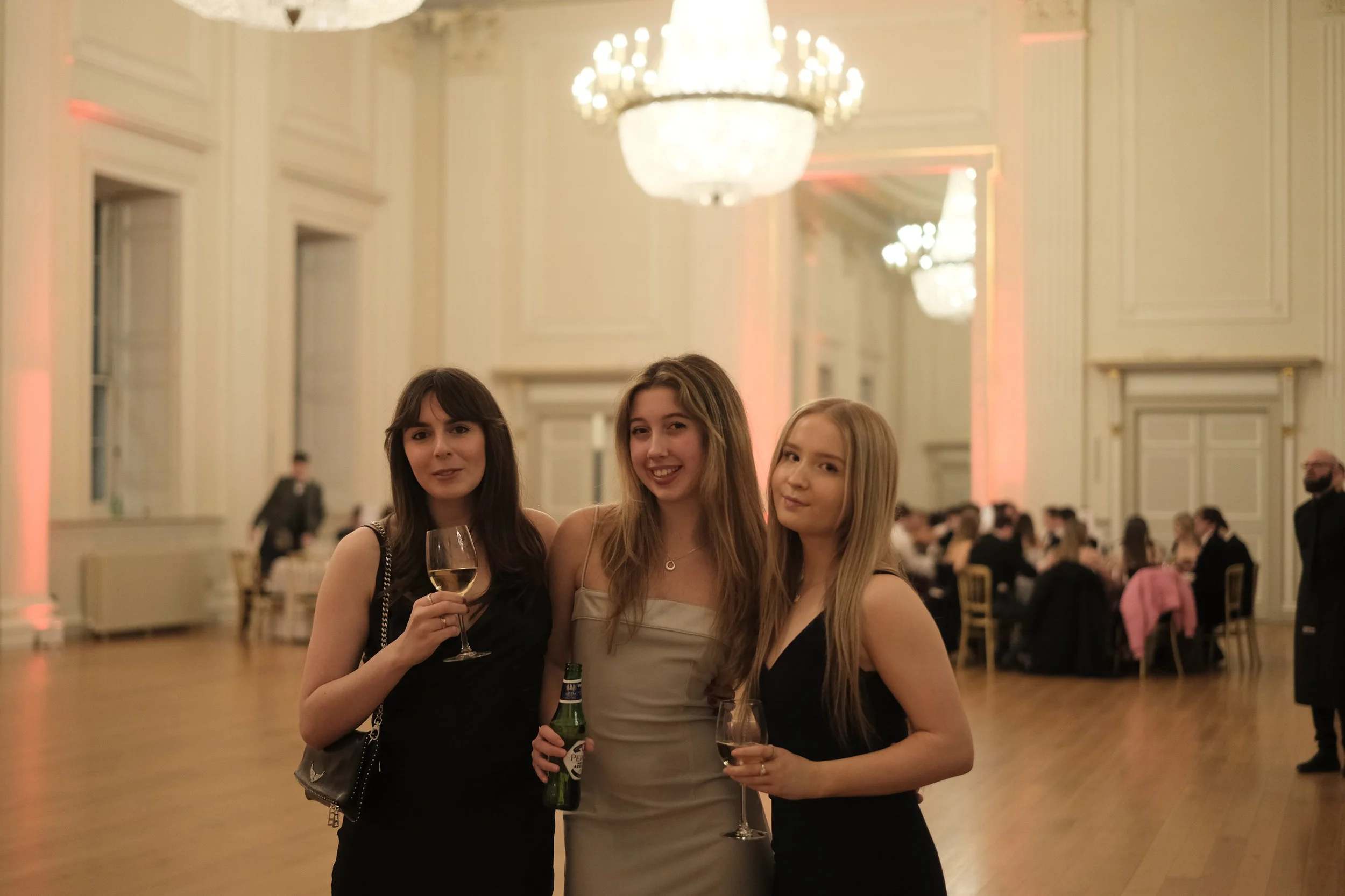 Three young women in formal attire standing in a ballroom with chandeliers, holding wine glasses.
