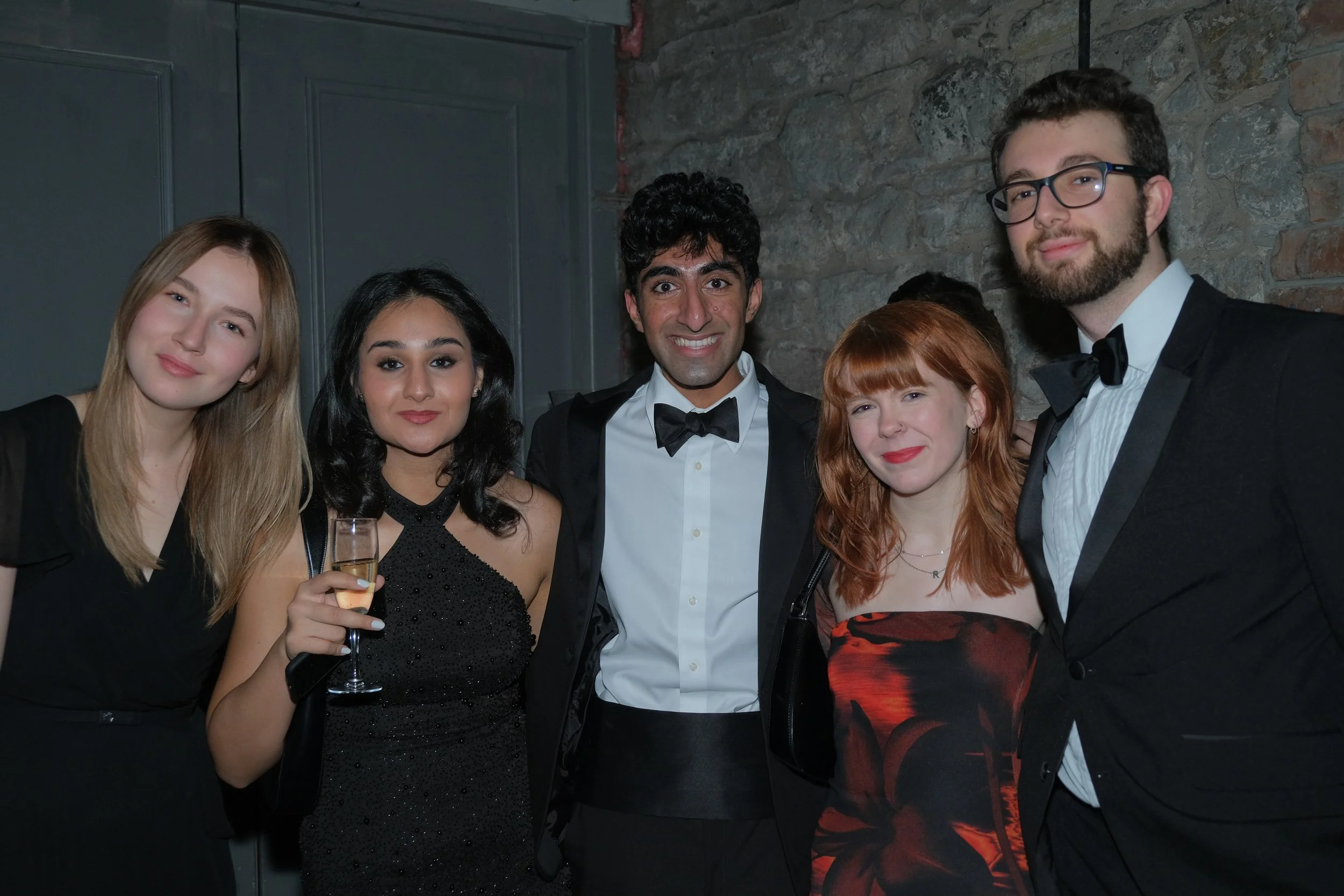Group of five young adults dressed in formal attire at a social event, smiling, with one holding a glass of champagne.