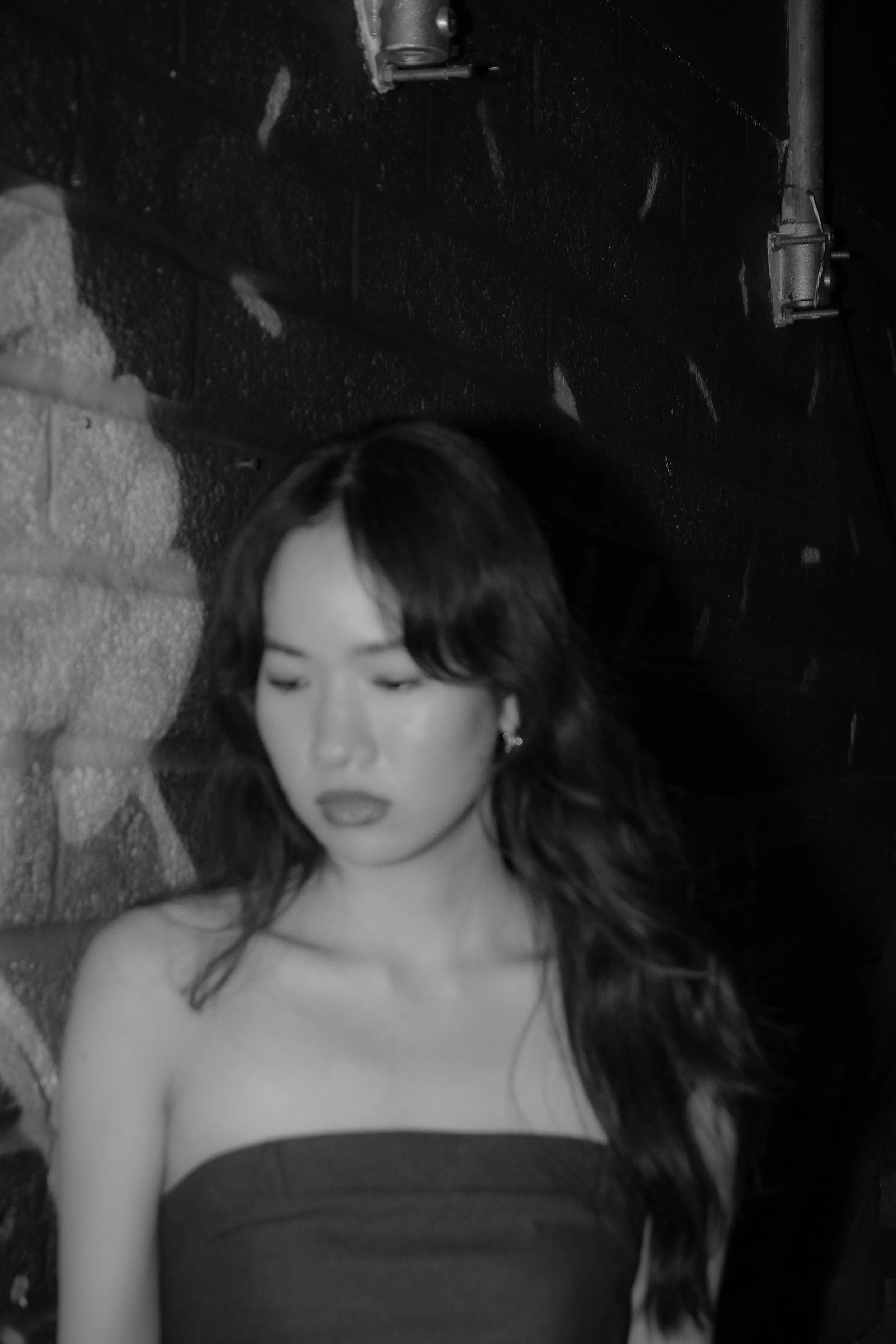 A black and white photo of a woman with long hair, wearing a strapless top, with her eyes closed, leaning against a brick wall.