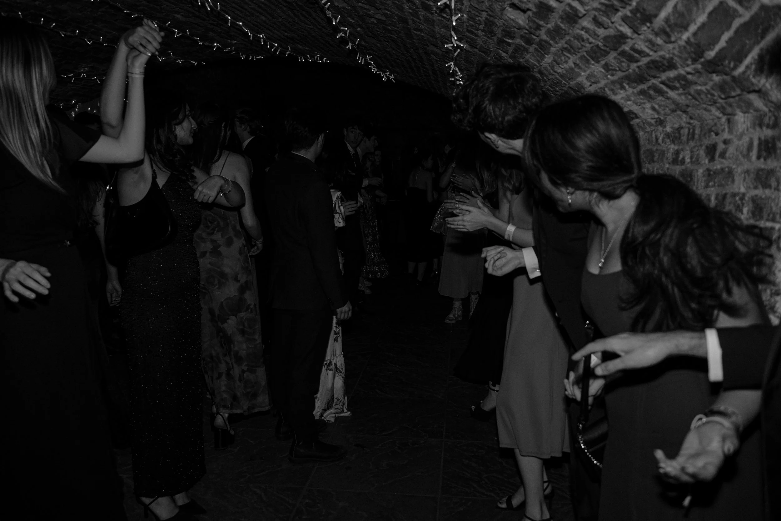 People dancing and socializing at a party in a dimly lit, brick-walled venue with string lights overhead.