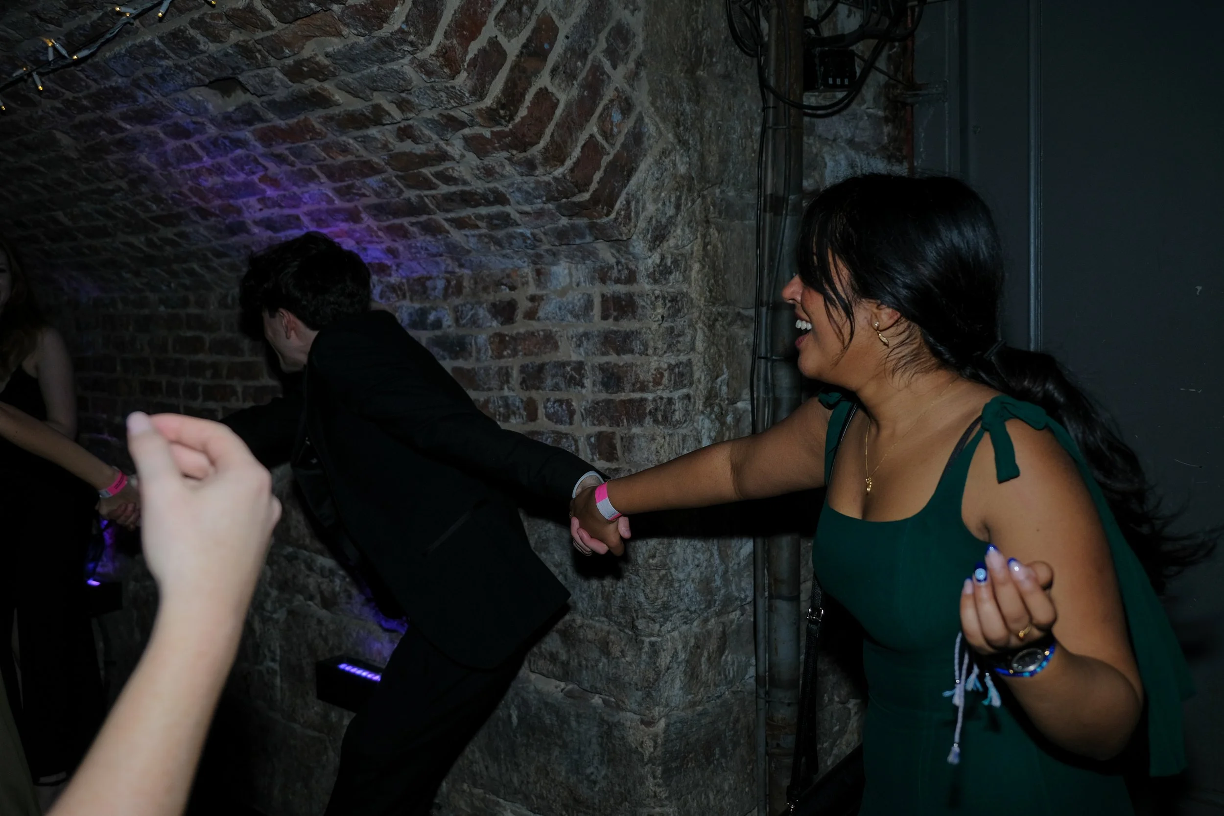 A woman in a green dress is holding hands with a man in a black suit and they are dancing or playing in a dimly lit space with brick walls and purple lighting.
