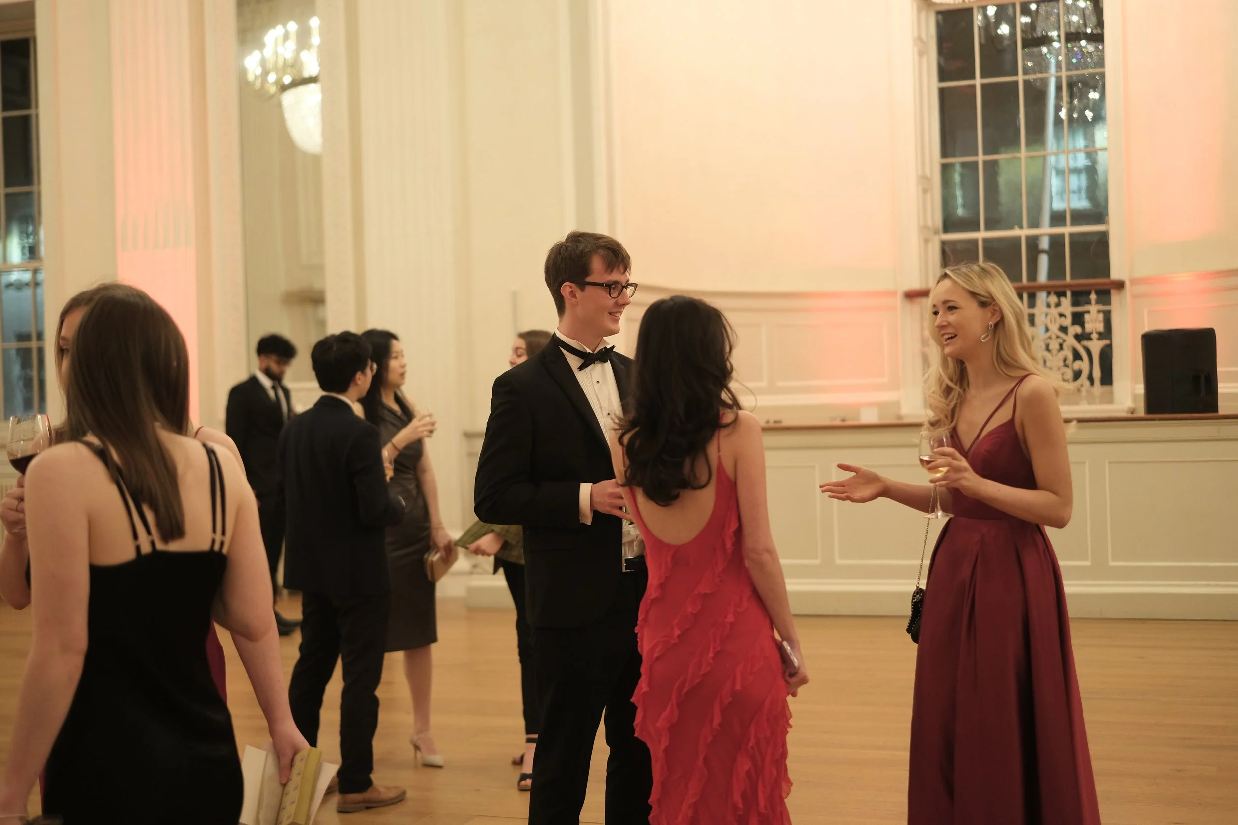 People dressed in formal wear, socializing in an elegant ballroom with large windows and ornate decorations.