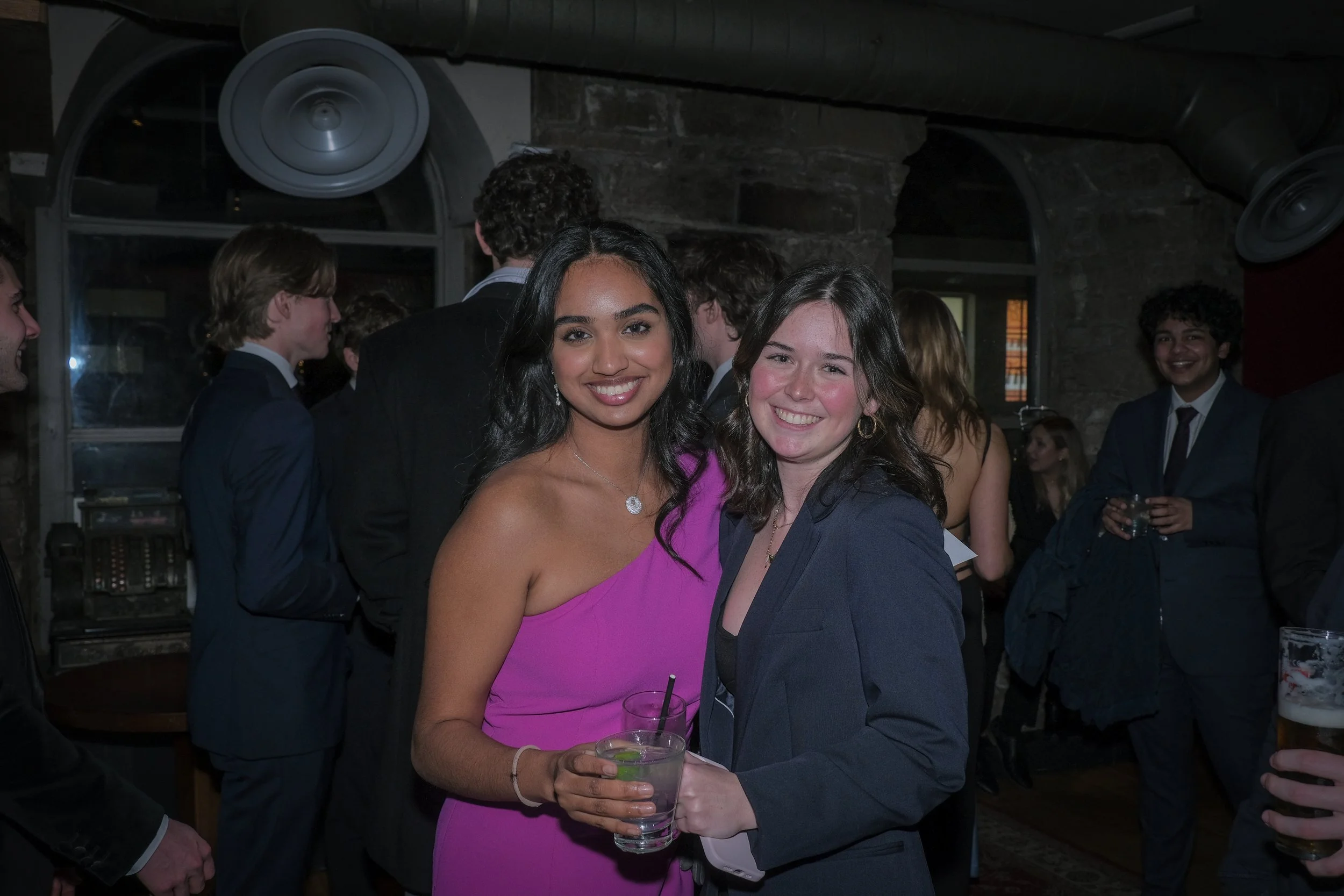 Two young women smiling and holding drinks at a social event, with other attendees mingling in the background.