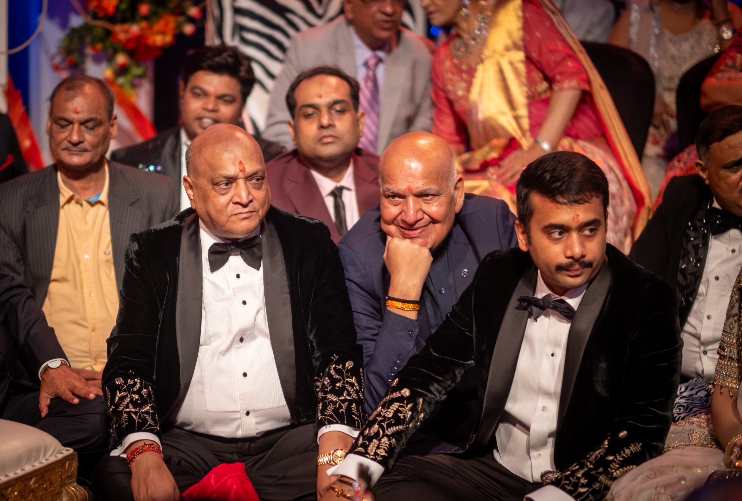 Group of men and women dressed in formal and traditional Indian attire, sitting at a celebration or wedding event, with colorful decorations in the background.