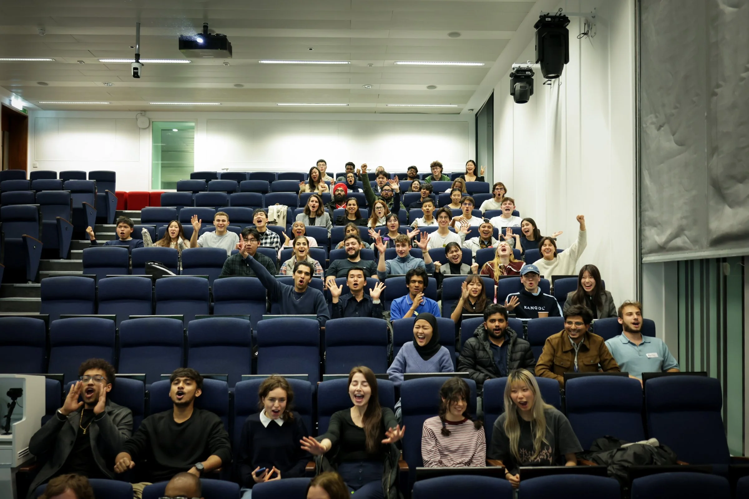 A diverse group of young people in a lecture hall or conference room, smiling, cheering, and raising their hands, with some making peace signs, in an enthusiastic atmosphere.