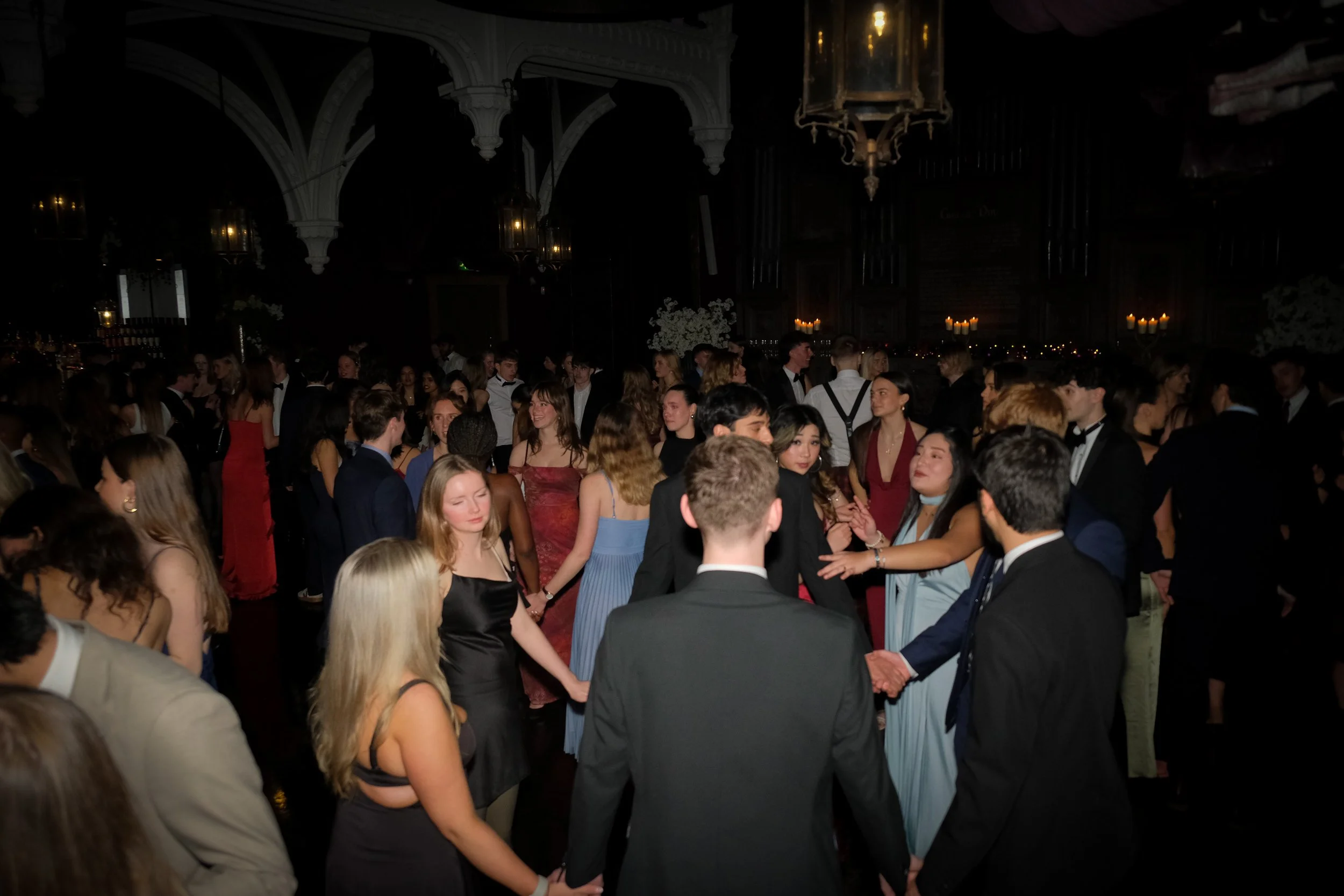 Crowd of young people dancing at a formal event in a large, dark hall with chandeliers and candlelight decorations.