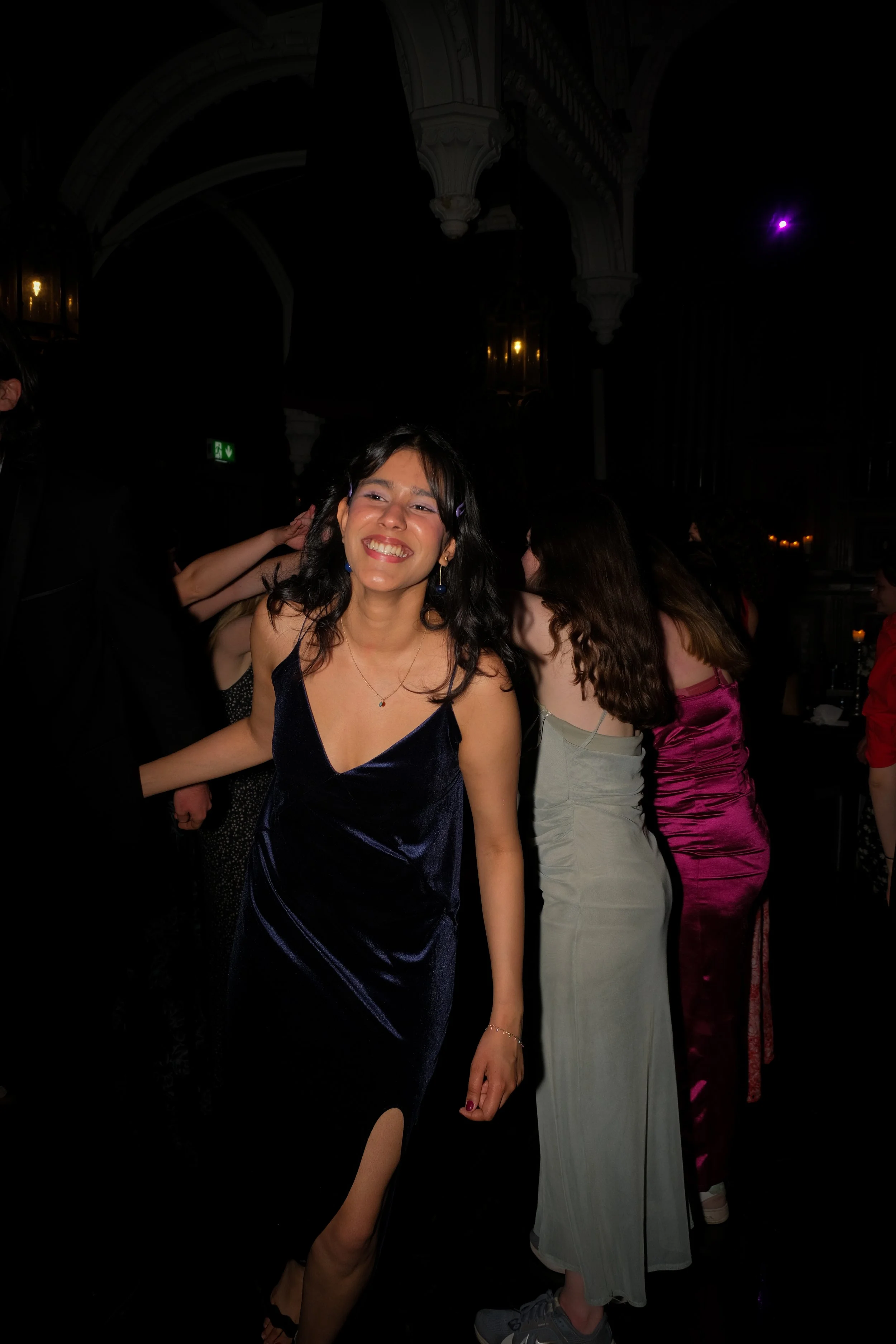 A woman in a dark blue dress smiling at a social event, with other women in colorful dresses behind her.