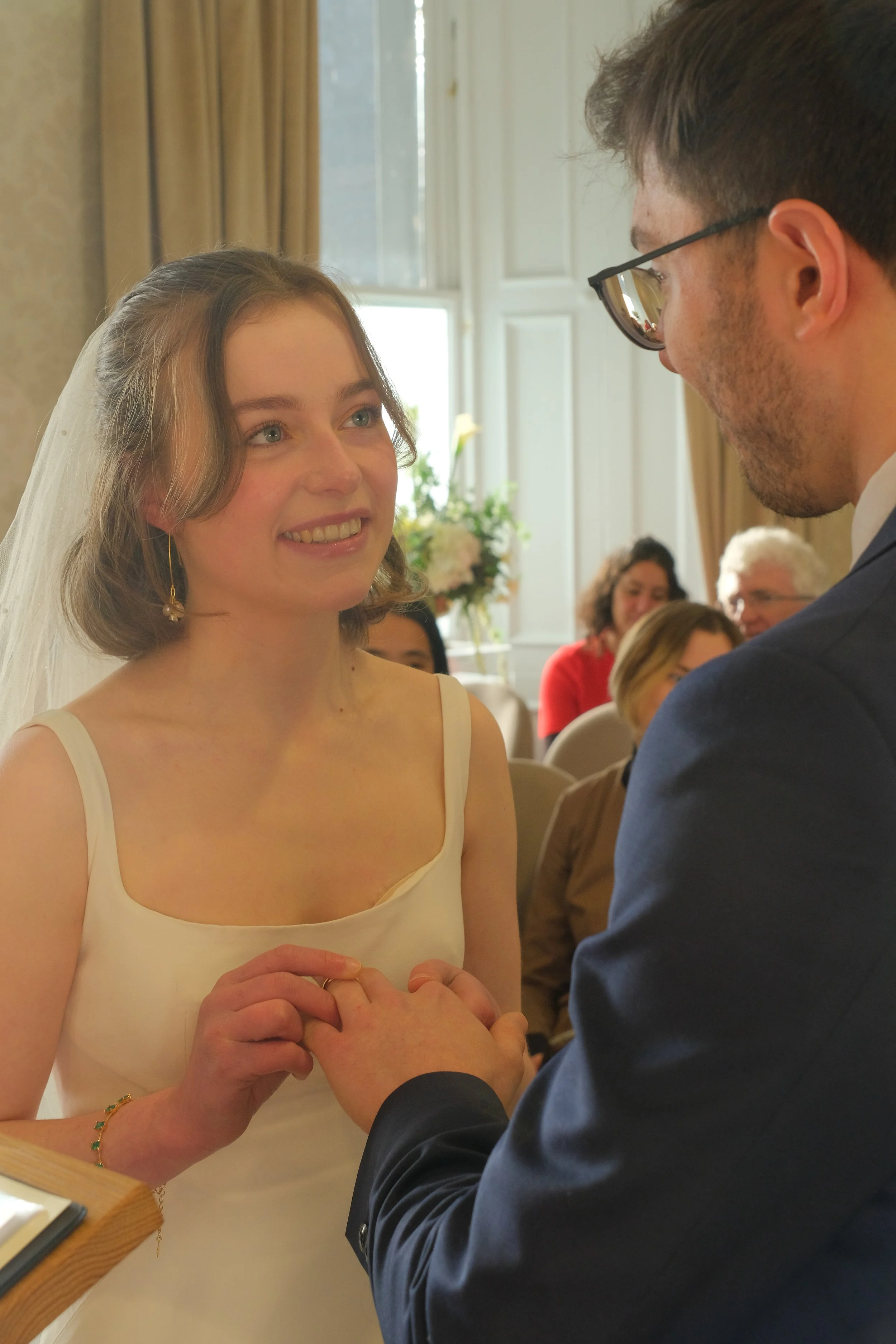 A bride and groom exchanging wedding vows, holding hands during their wedding ceremony in a well-lit room with guests watching.