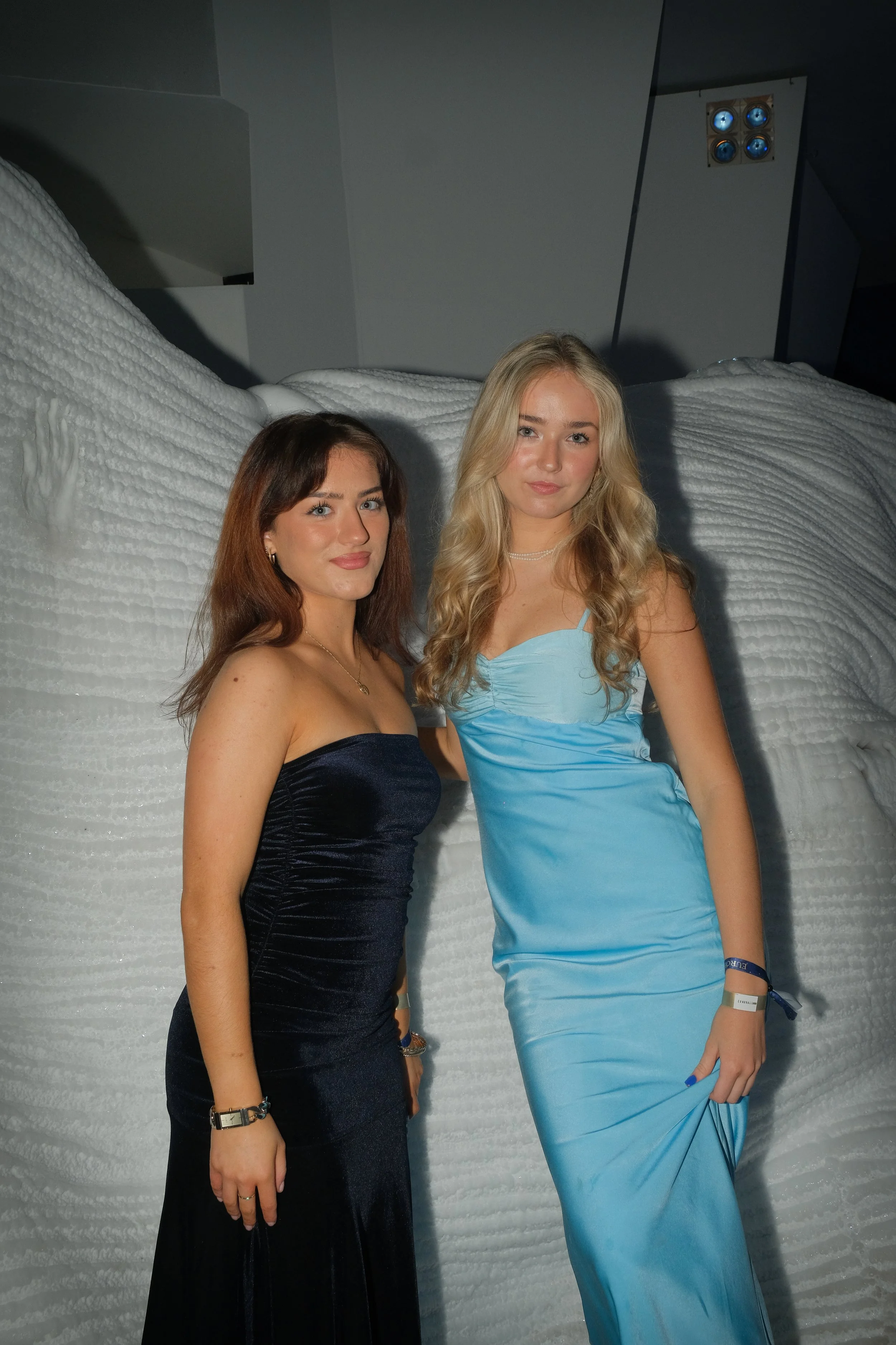Two young women in elegant dresses standing in front of a white textured surface with a grey wall and some lights in the background.