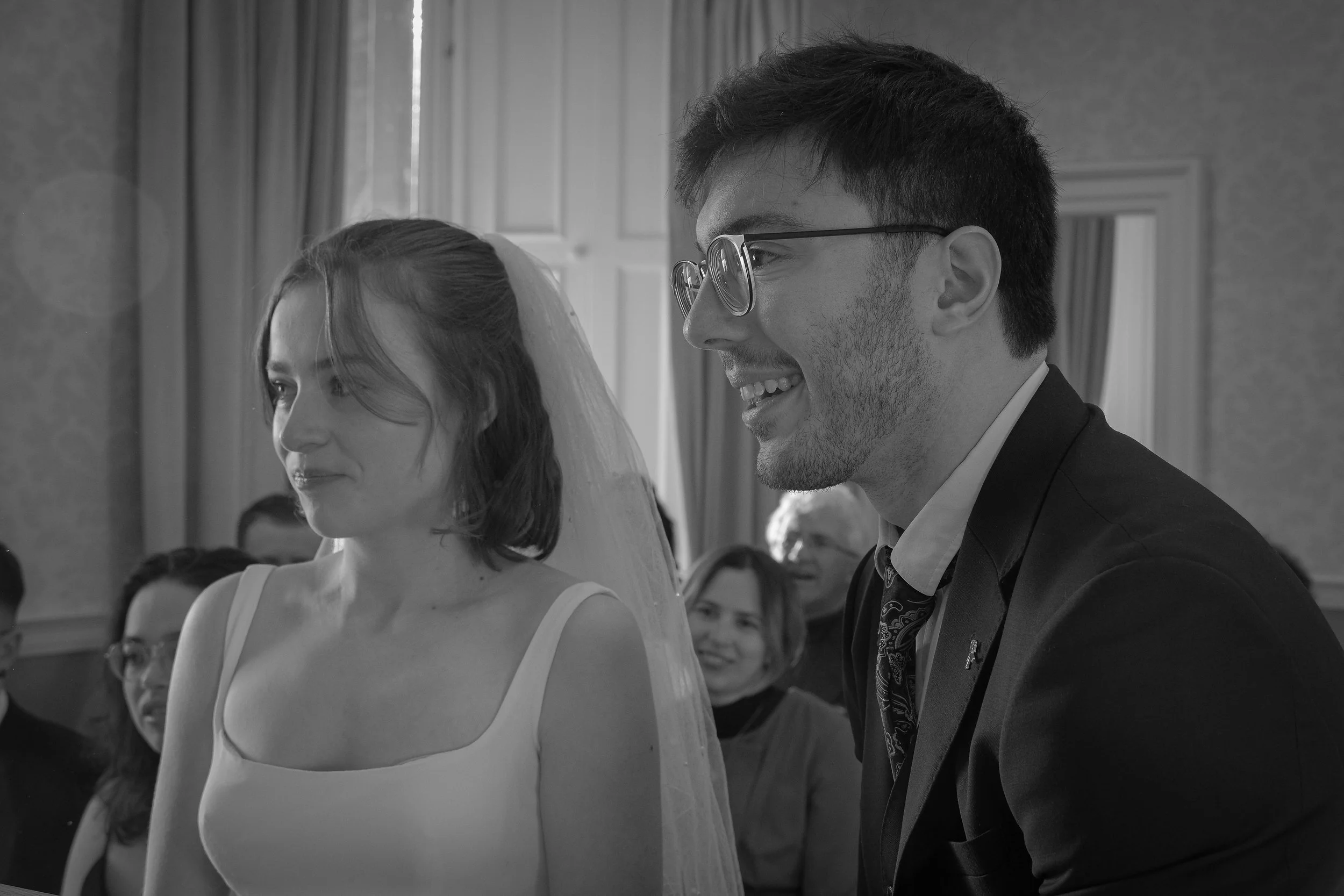 A black-and-white photo of a wedding ceremony showing a smiling groom in glasses and a bride with shoulder-length hair and a veil, surrounded by seated guests.