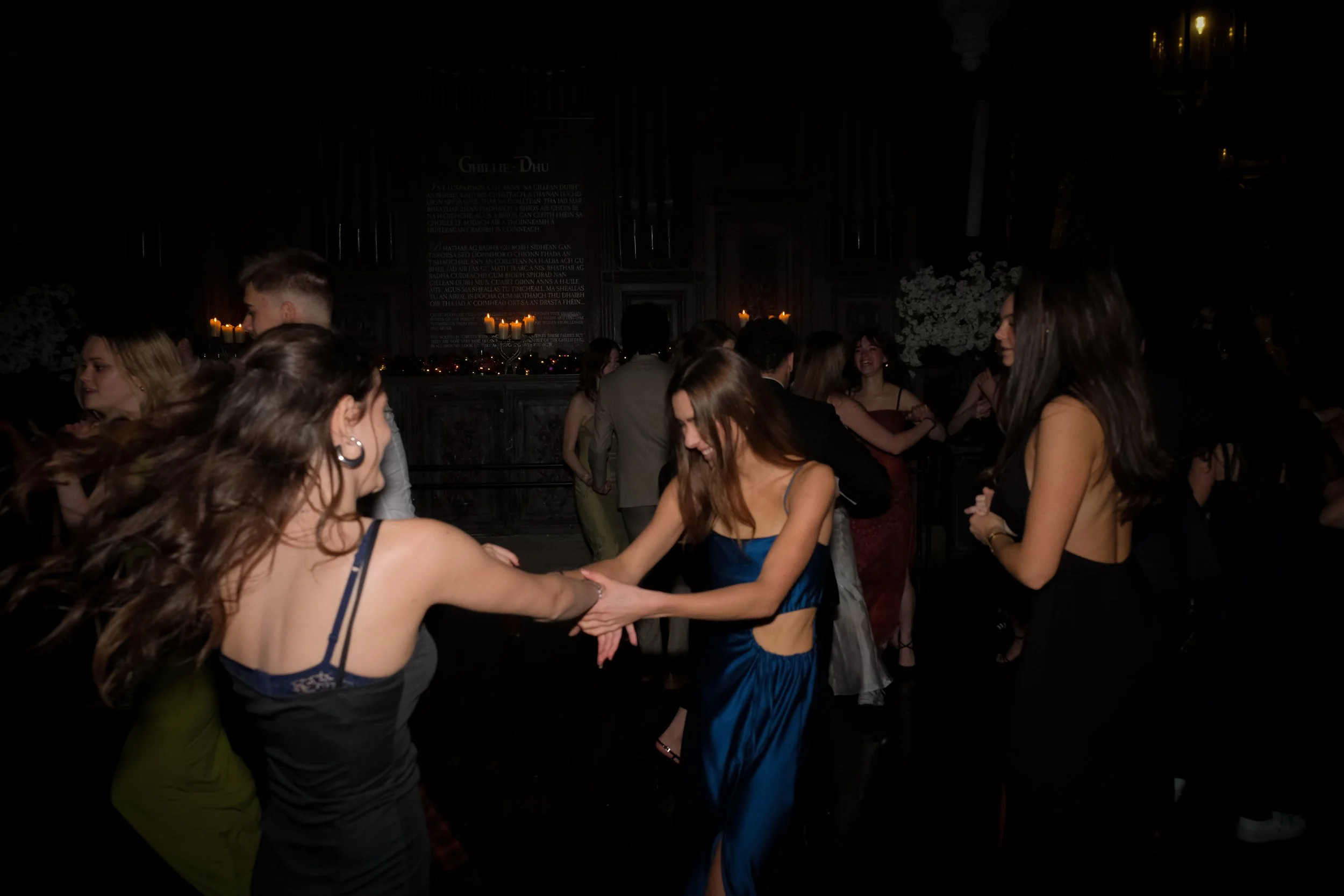 People dancing and socializing at a dark indoor event, some women are holding hands and smiling, dressed in formal evening wear.