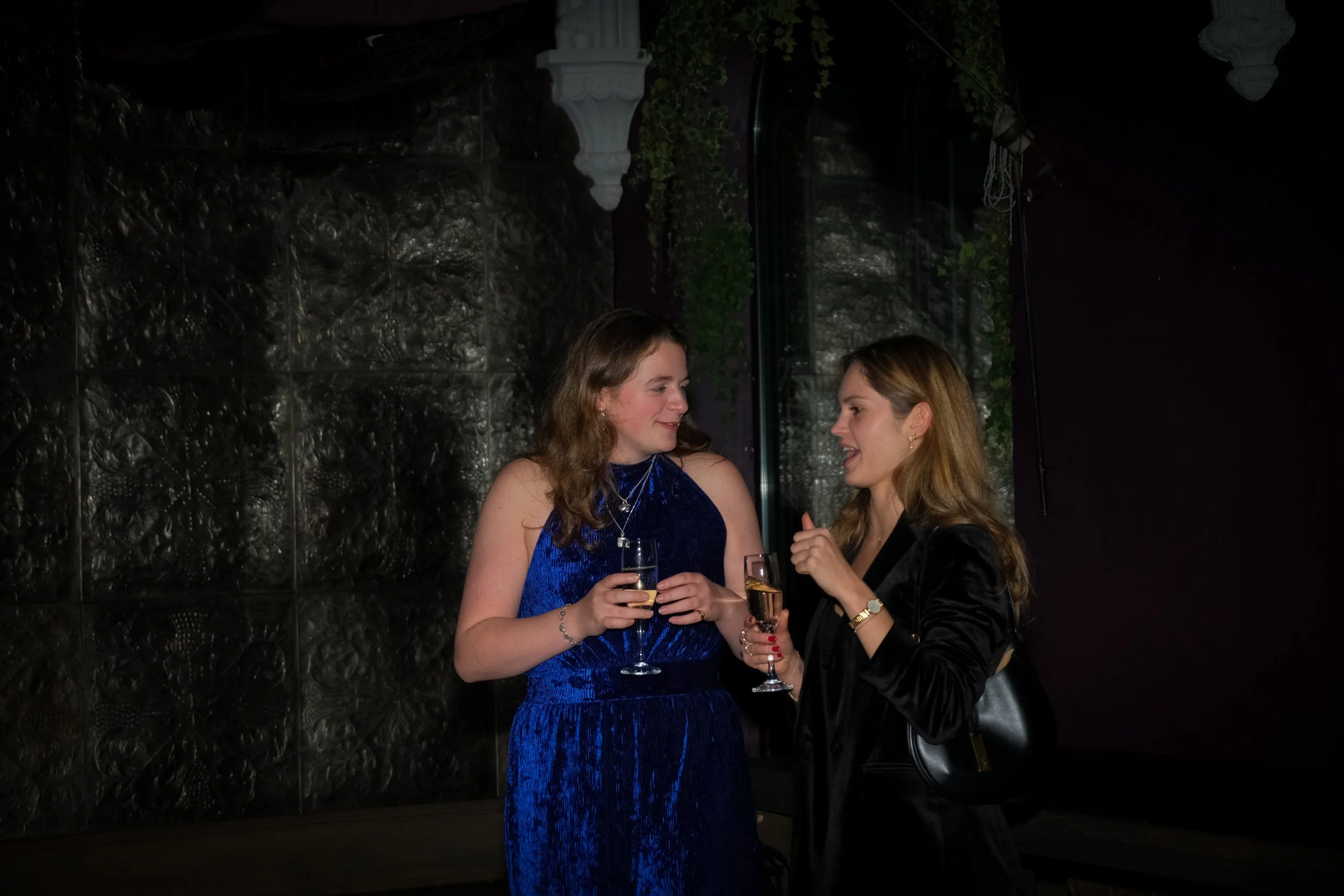 Two women with light skin and long hair are talking and holding champagne glasses in a dimly lit room. One woman is wearing a blue dress, and the other is wearing a black outfit.