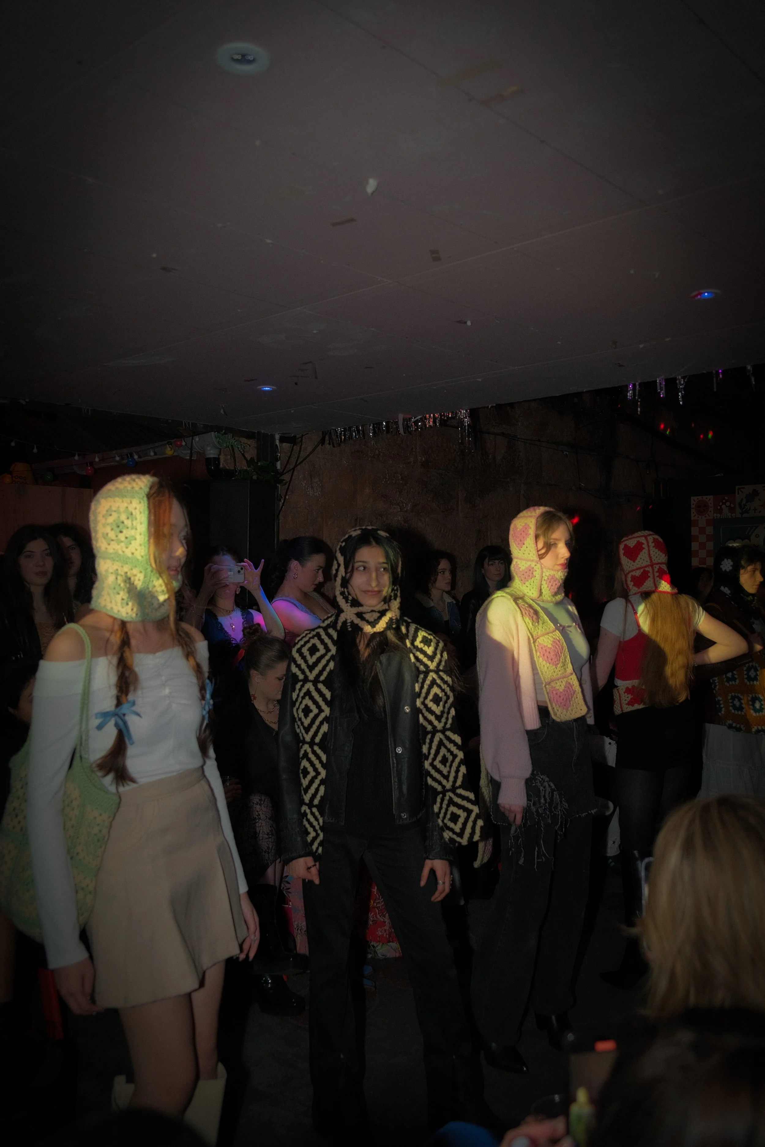 A group of women standing on a stage or runway at a fashion event or party. Some women are wearing knitted masks with colorful patterns, and there are multiple people in the background.