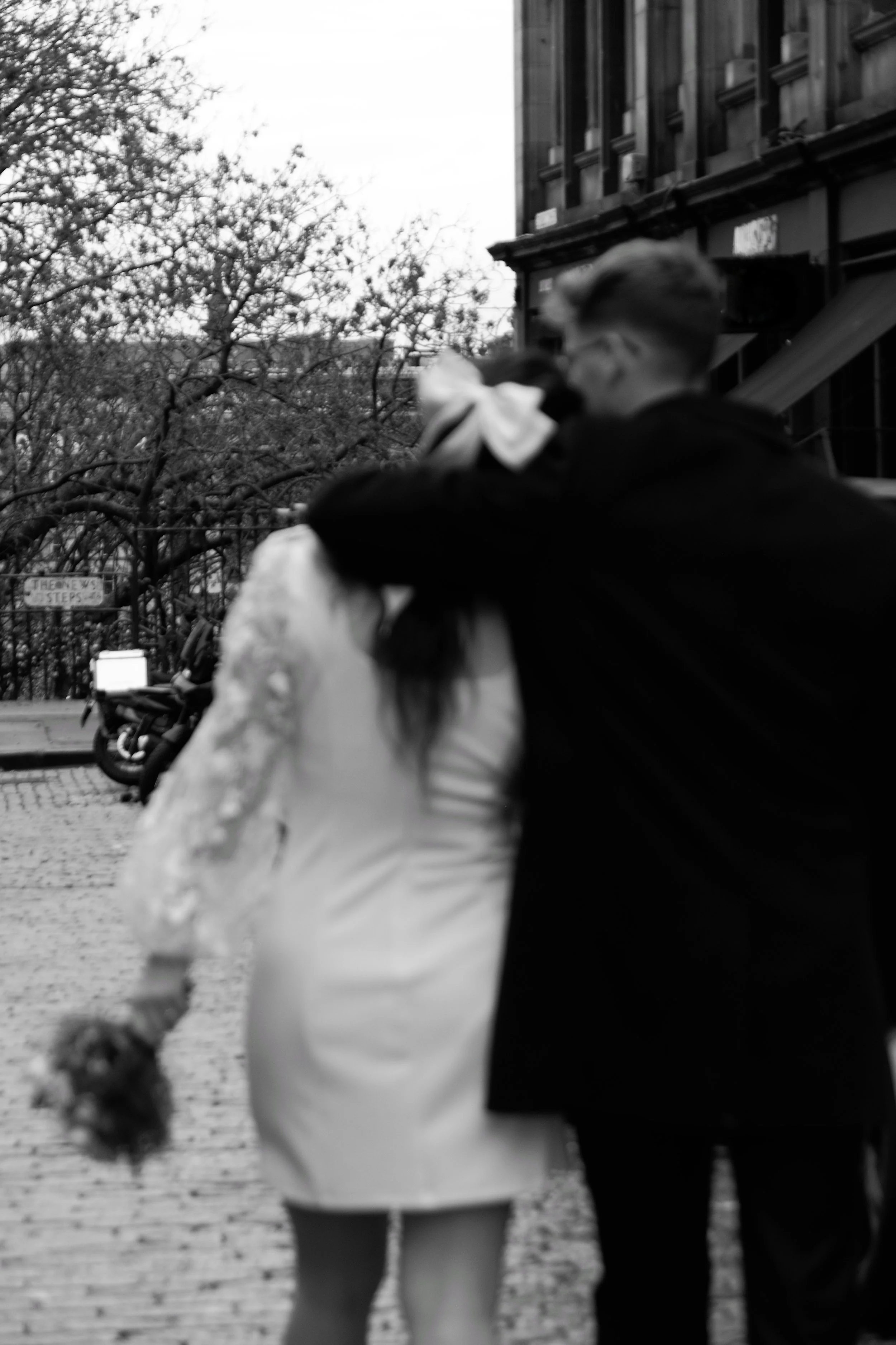 A black-and-white photo of a couple walking together on a city sidewalk, with the person on the left holding a bouquet of flowers, and the person on the right embracing them. The background shows a building, trees, parked motorcycles, and a street si